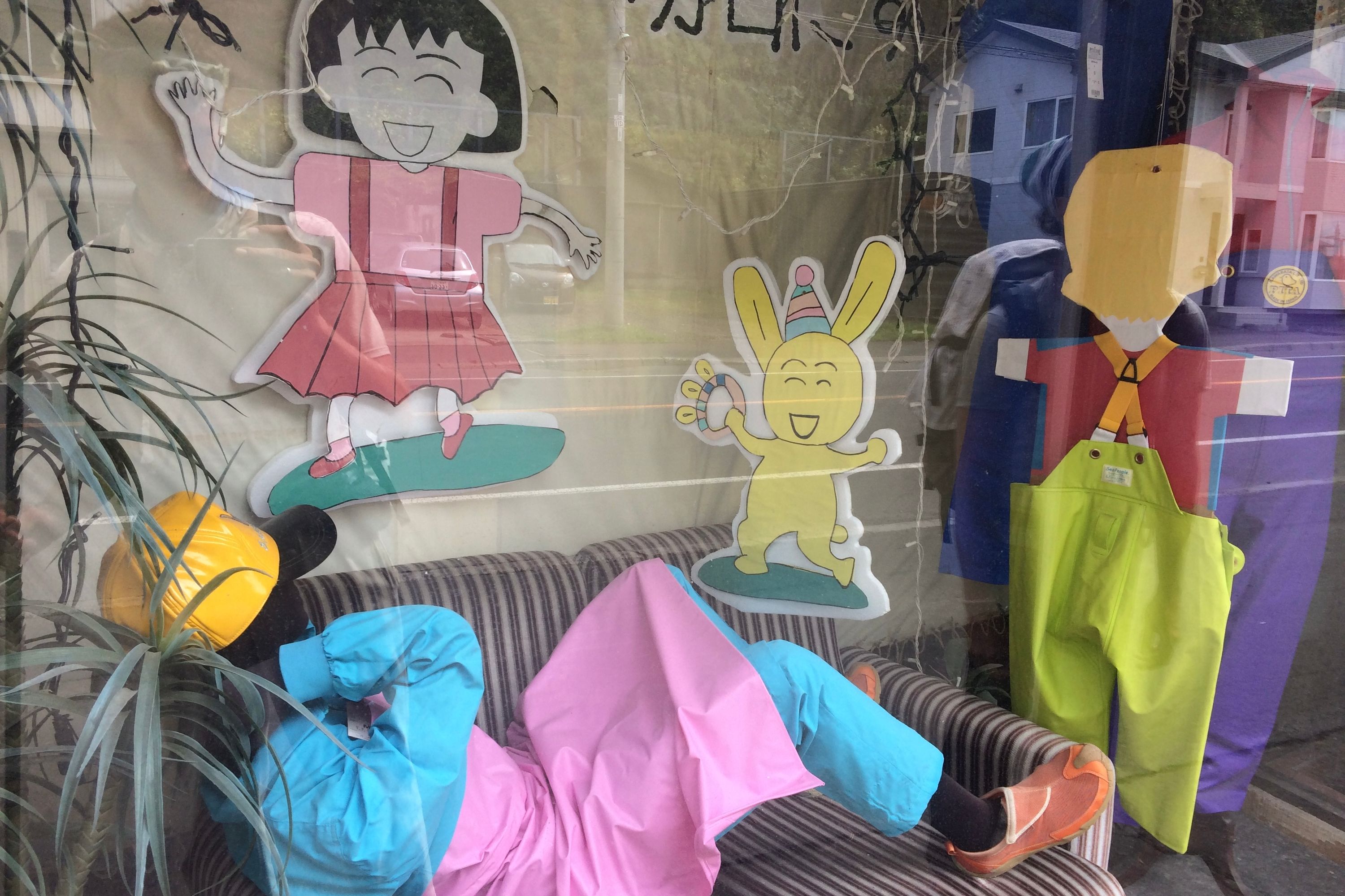 A person in colorful clothes rests among other colorful things in the window display of a shop.