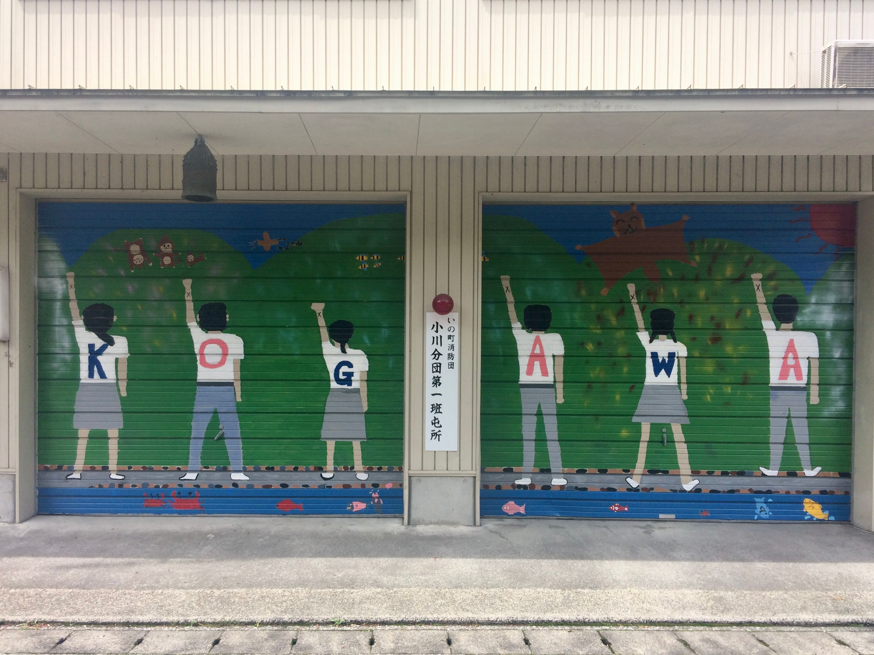 A giant flying squirrel makes a low pass over six children on a garage door mural, their left arms raised to the sky with clenched fists, the backs of their white shirts spelling “KOGAWA”.