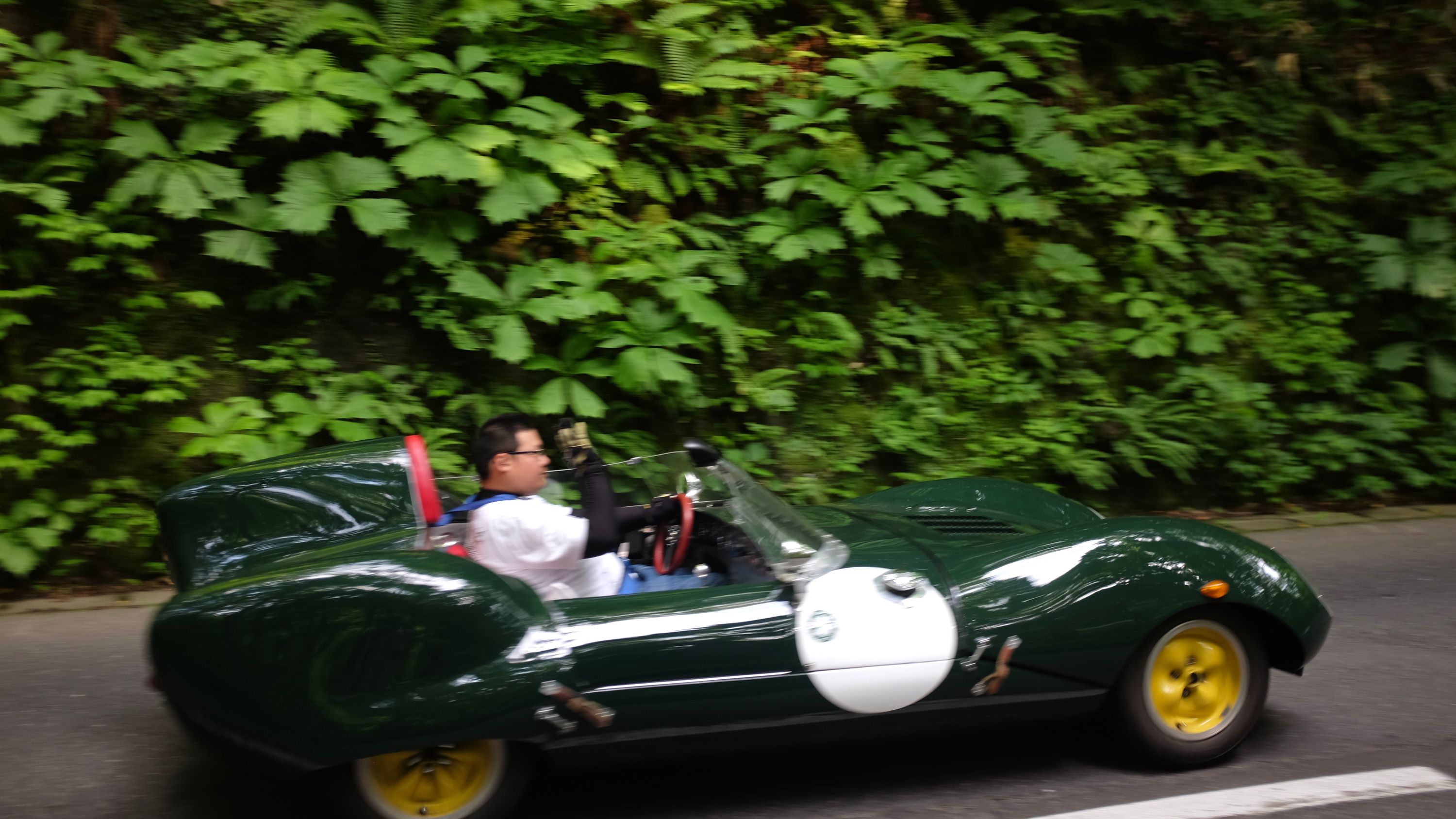 A man drives a low-slung green sports car, a Lotus Eleven, on a forest road.