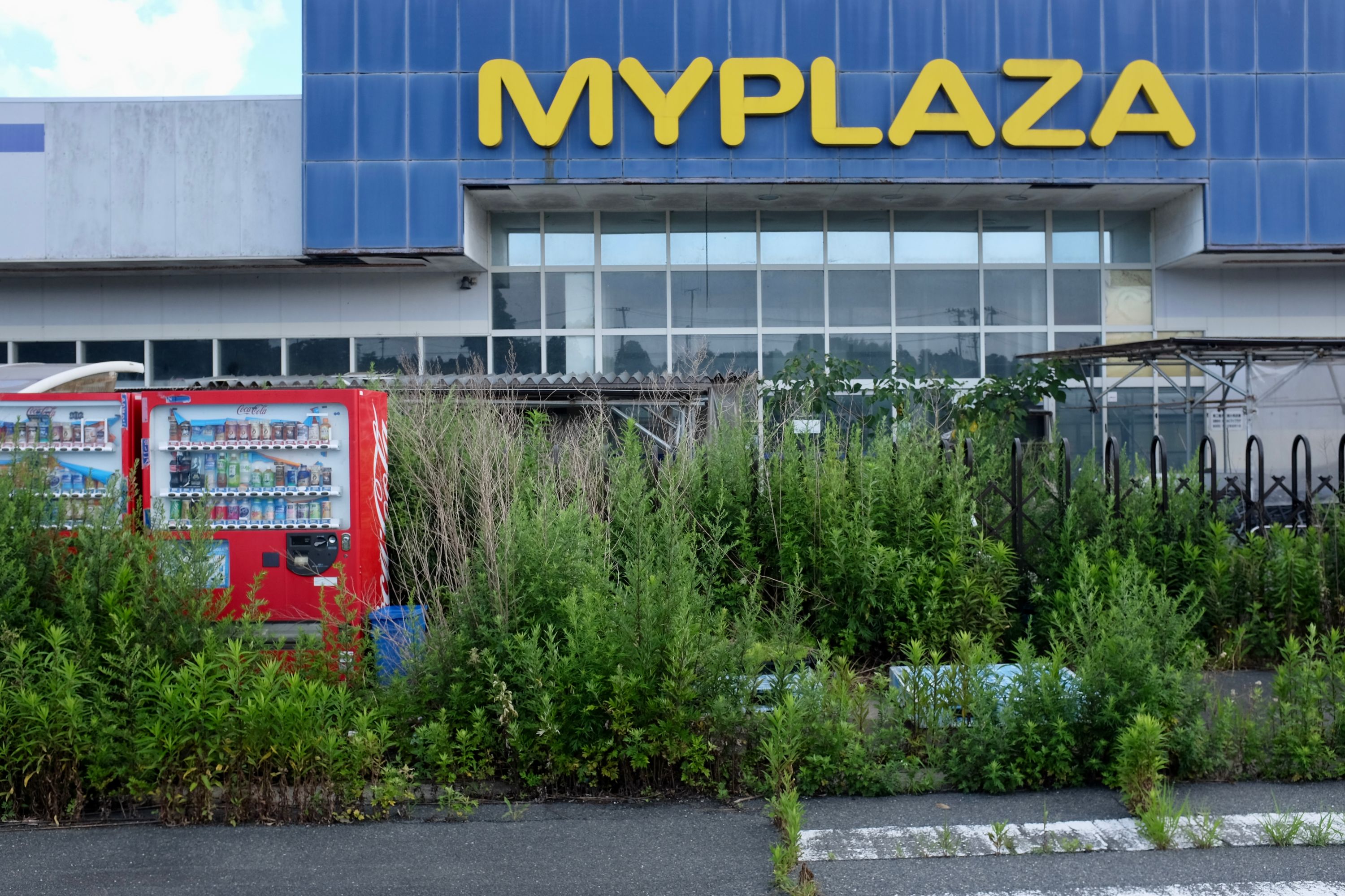 Vending machines overgrown with weeds in front of an abandoned shopping center named Myplaza.
