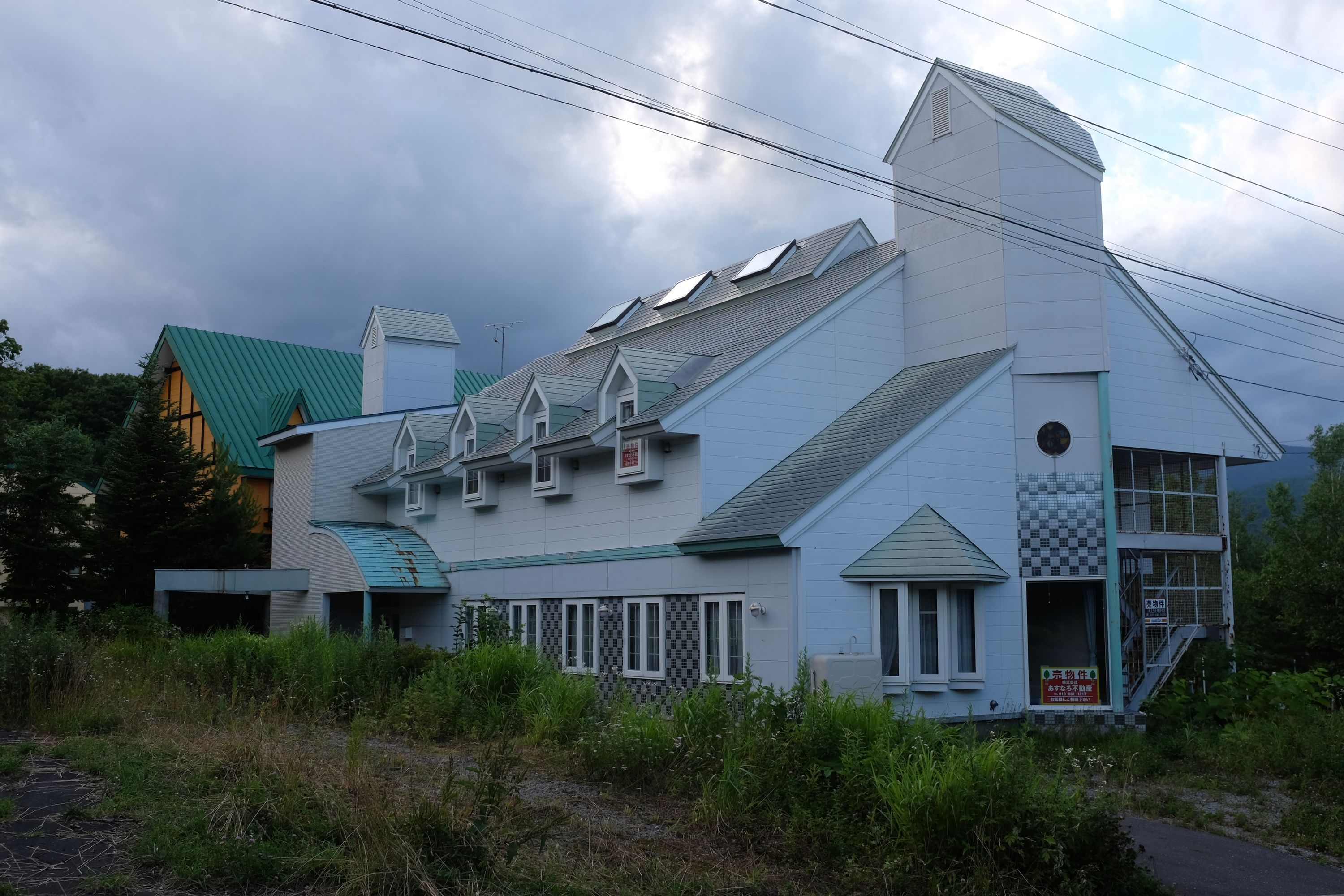 A very 90s building, white and shaped like a child’s drawing of a European church, lies abandoned in the weeds.