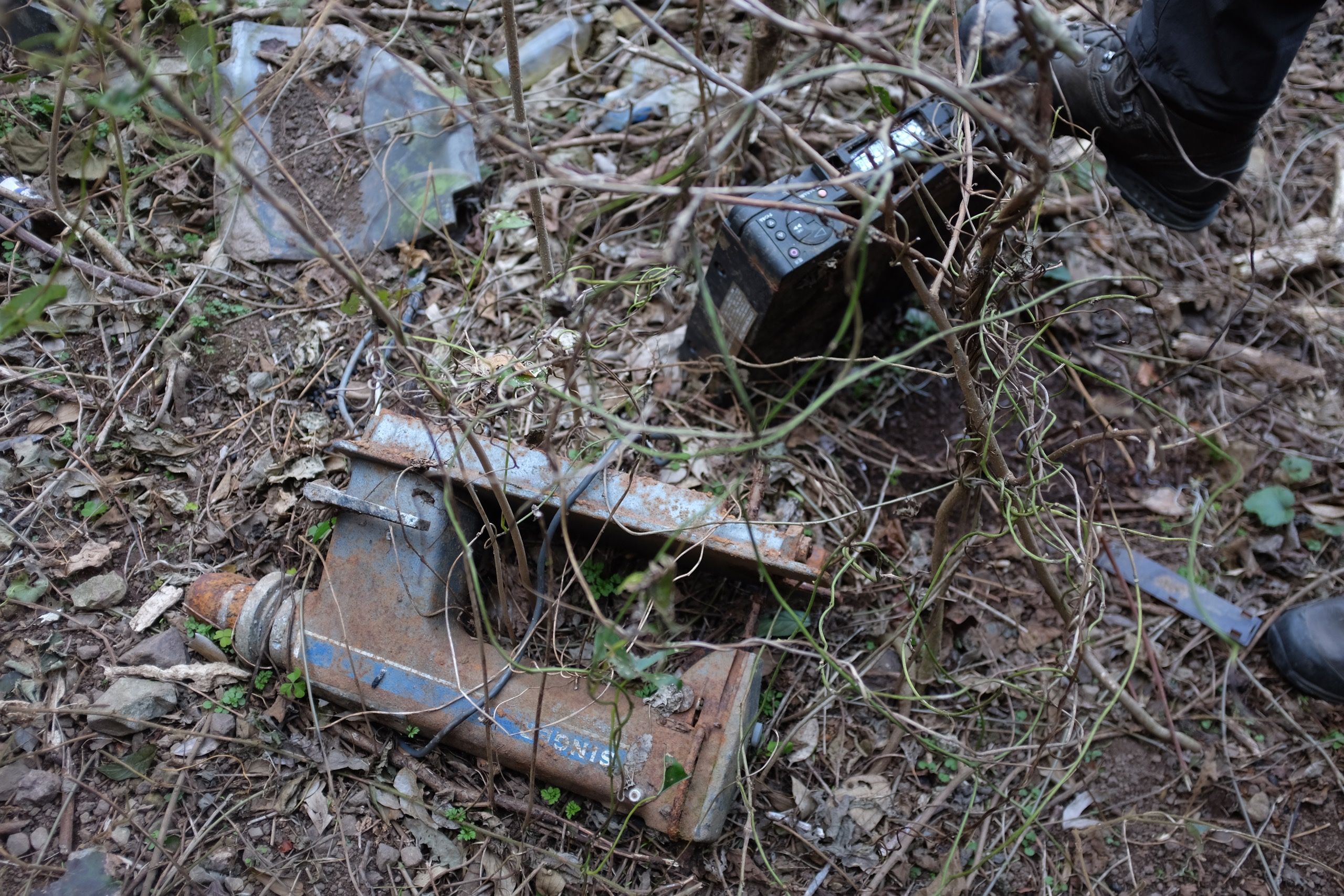 A man’s hiking boot steps on a discarded VCR, which lies among other pieces of garbage.