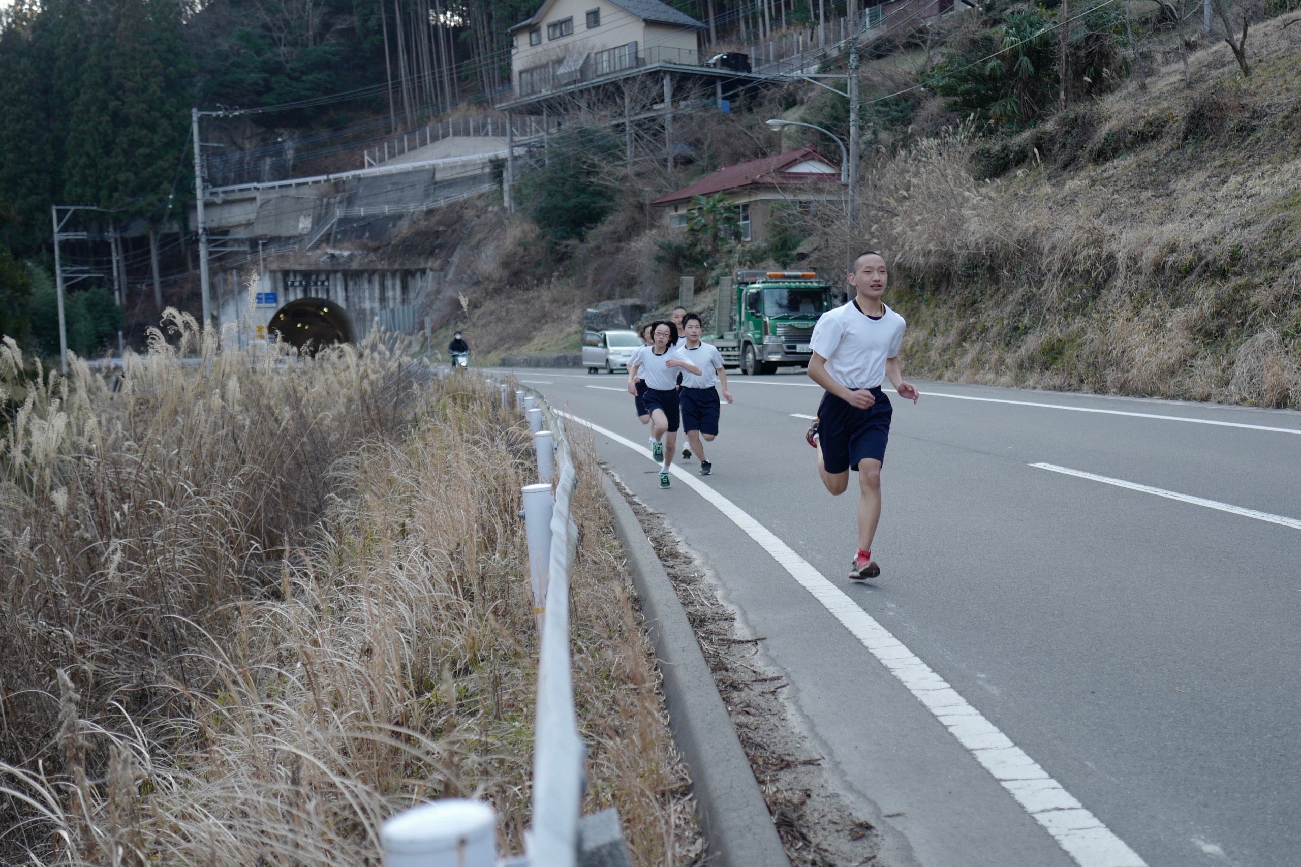 Schoolchildren in exercise clothes run along the side of a country road, with the entrance of a tunnel in the background.