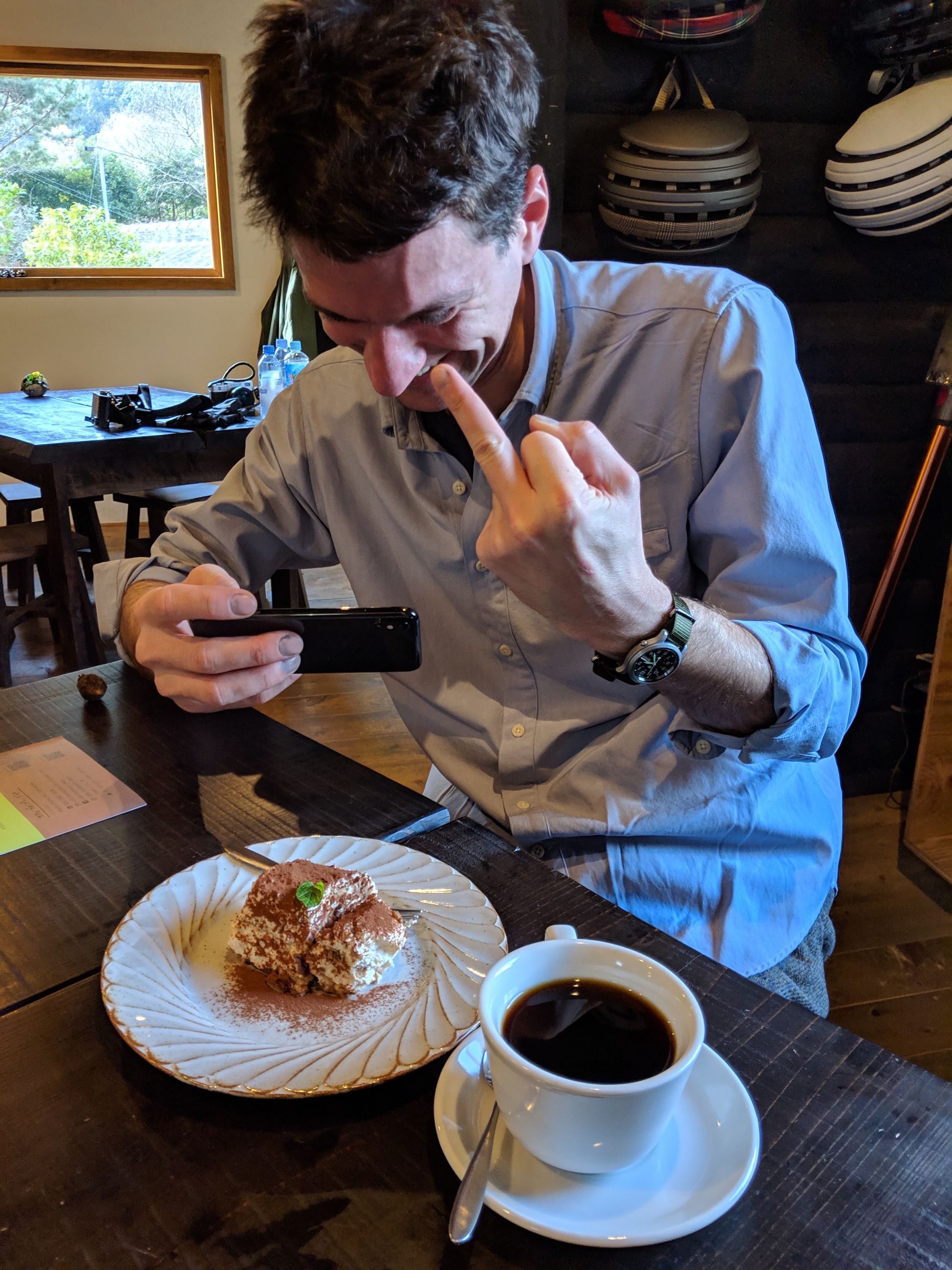 A man in a blue oxford shirt, the author, takes a photo of the cake with his cell phone while giving the finger to someone, presumably the photographer.
