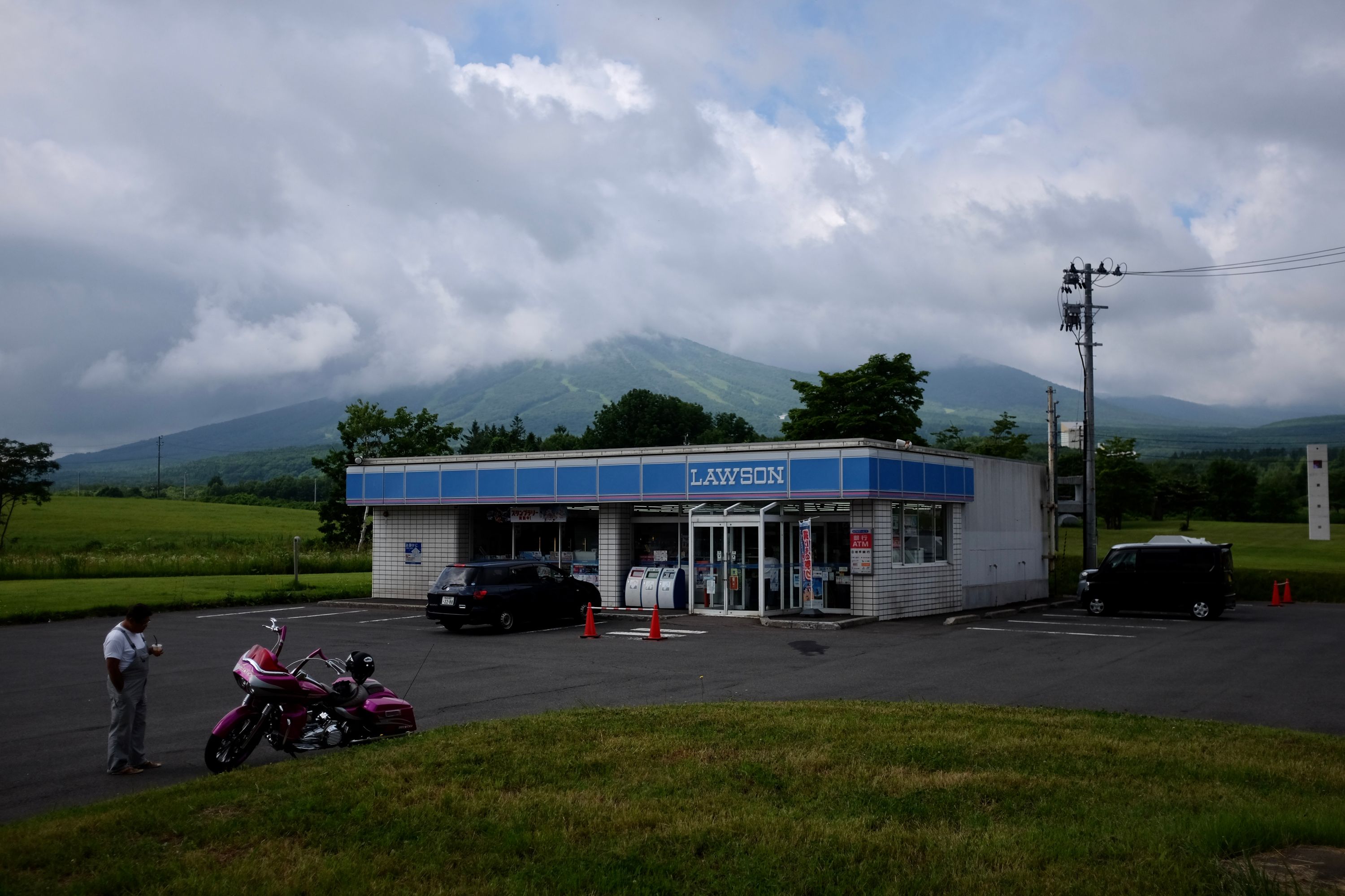 A Lawson convenience store on a cloudy day, with a forested mountain on the horizon.