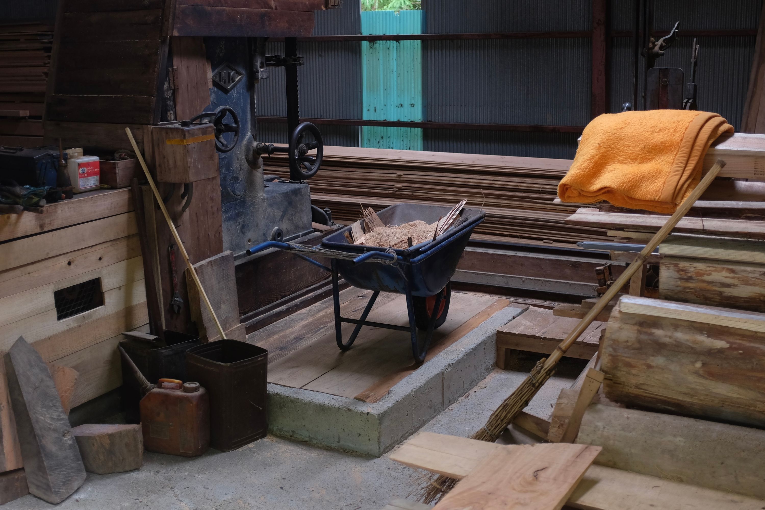The inside of a sawmill: planks, timber, wood-cutting machines, various tools, a blue wheelbarrow filled with sawdust and scraps.
