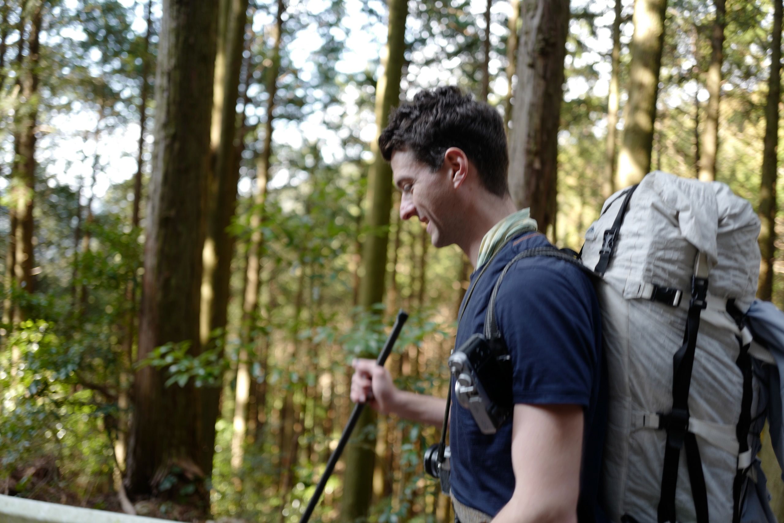 A color photograph of Peter Orosz at the beginning of his walking journey across Shikoku. He stands in a lush, green forest with tall trees creating a natural canopy overhead. Orosz wears a dark blue t-shirt and carries a large, light-colored hiking backpack with black straps. He holds a dark walking stick in his right hand. Dappled sunlight filters through the forest canopy, creating a serene, verdant atmosphere that suggests the peaceful start of a long trek through nature.