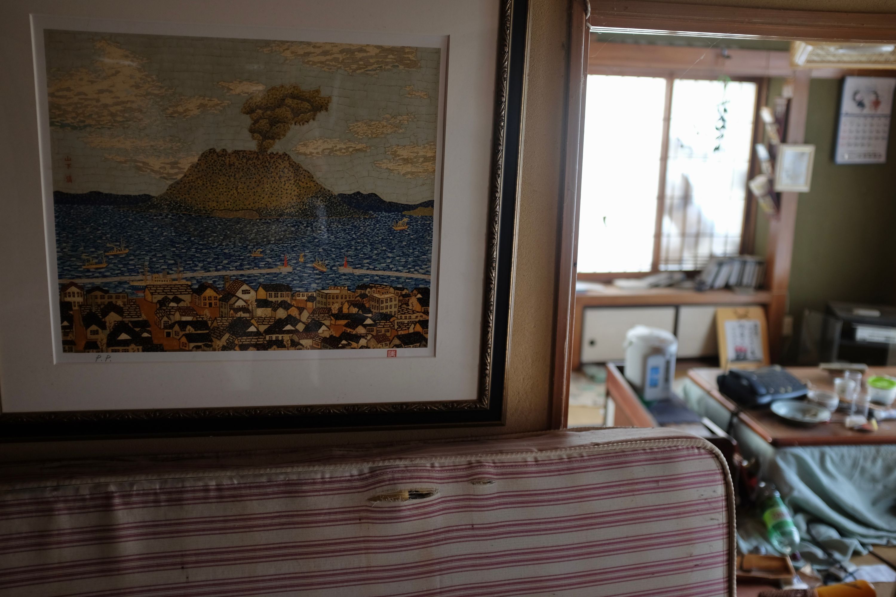 A painting of Kagoshima on the wall, and behind it a lot of scattered items in a kitchen.