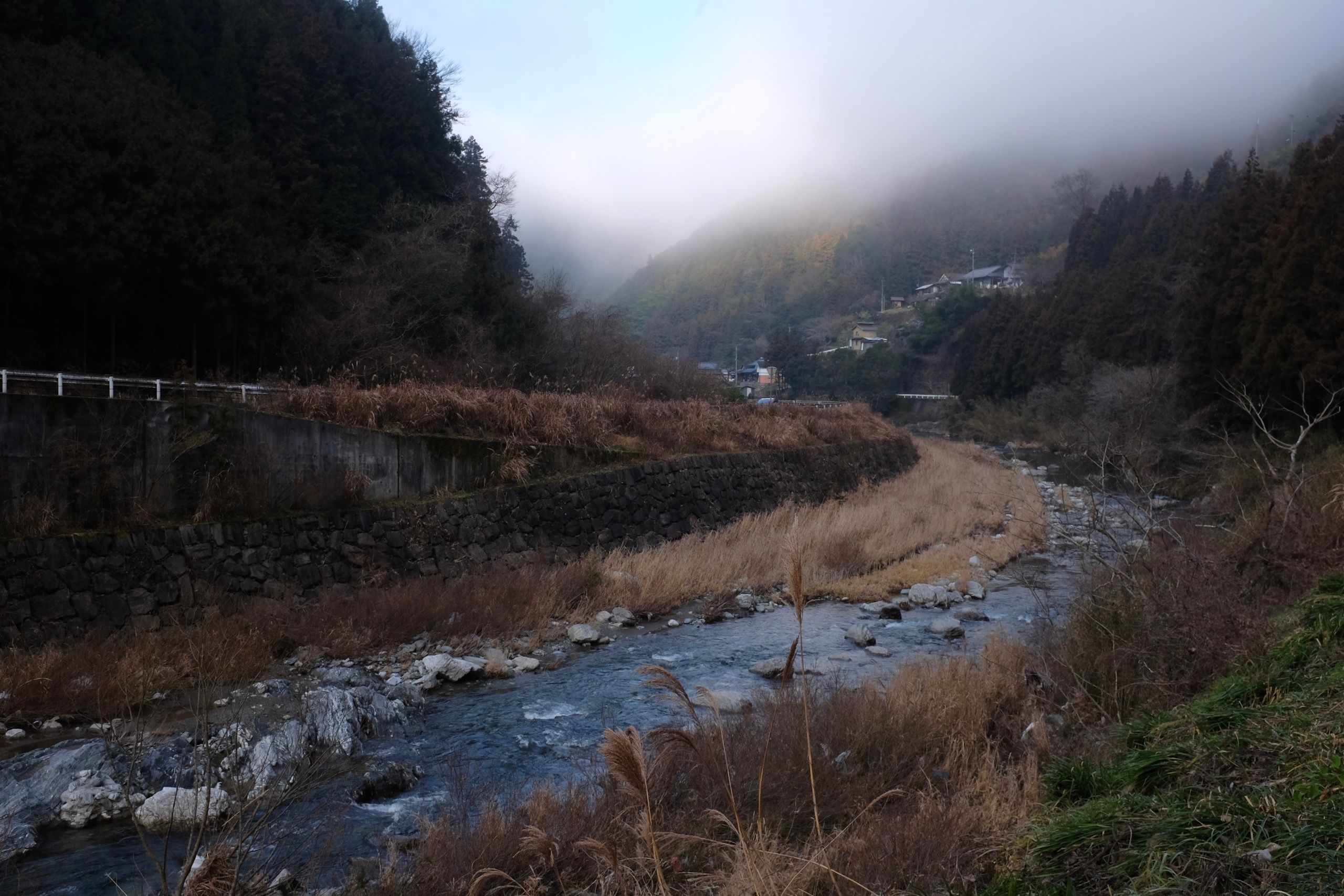 Fog lifts above a stream in a valley, with Japanese village houses on its banks in the distance.