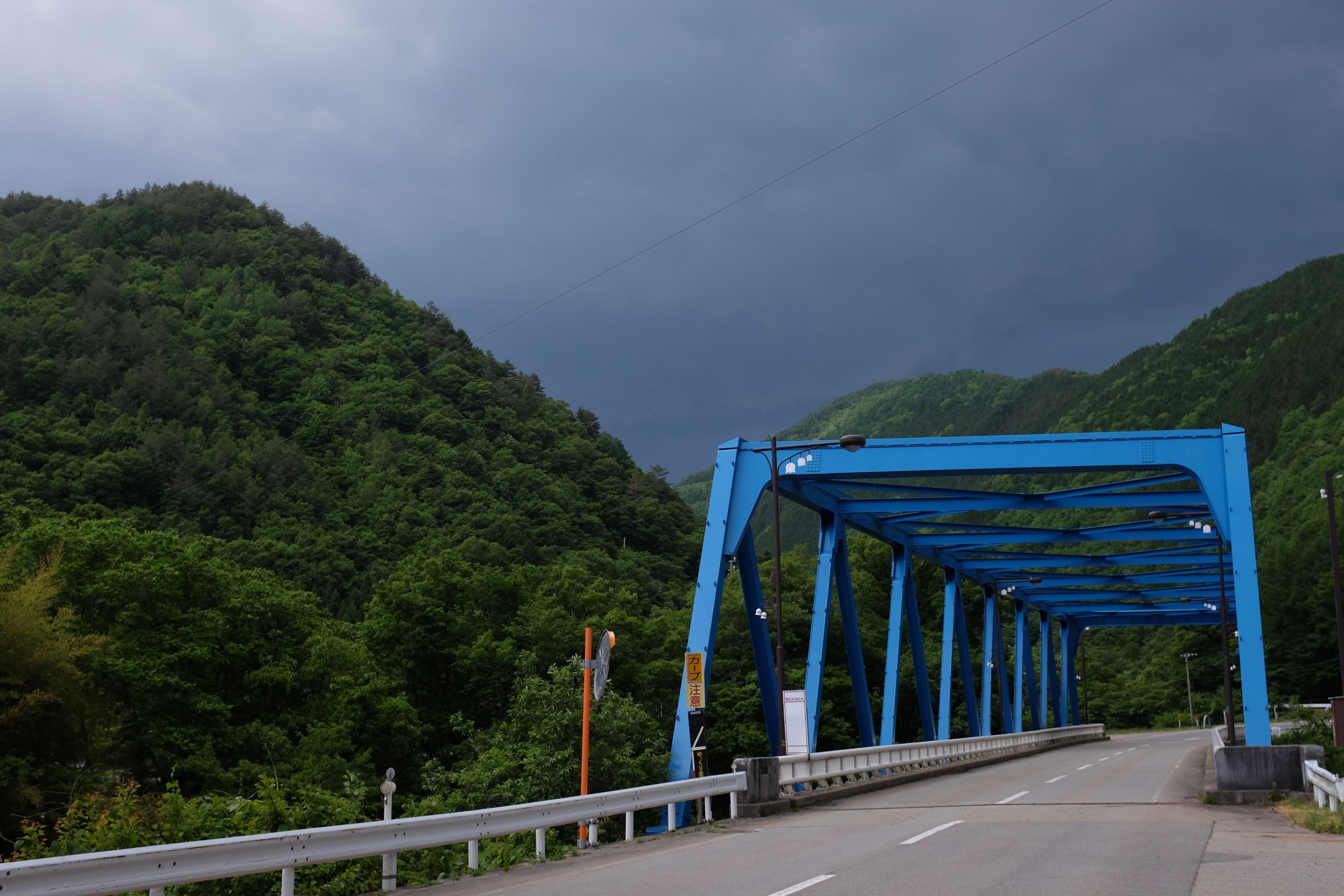 A blue bridge in front of forested hillsides and dark skies.