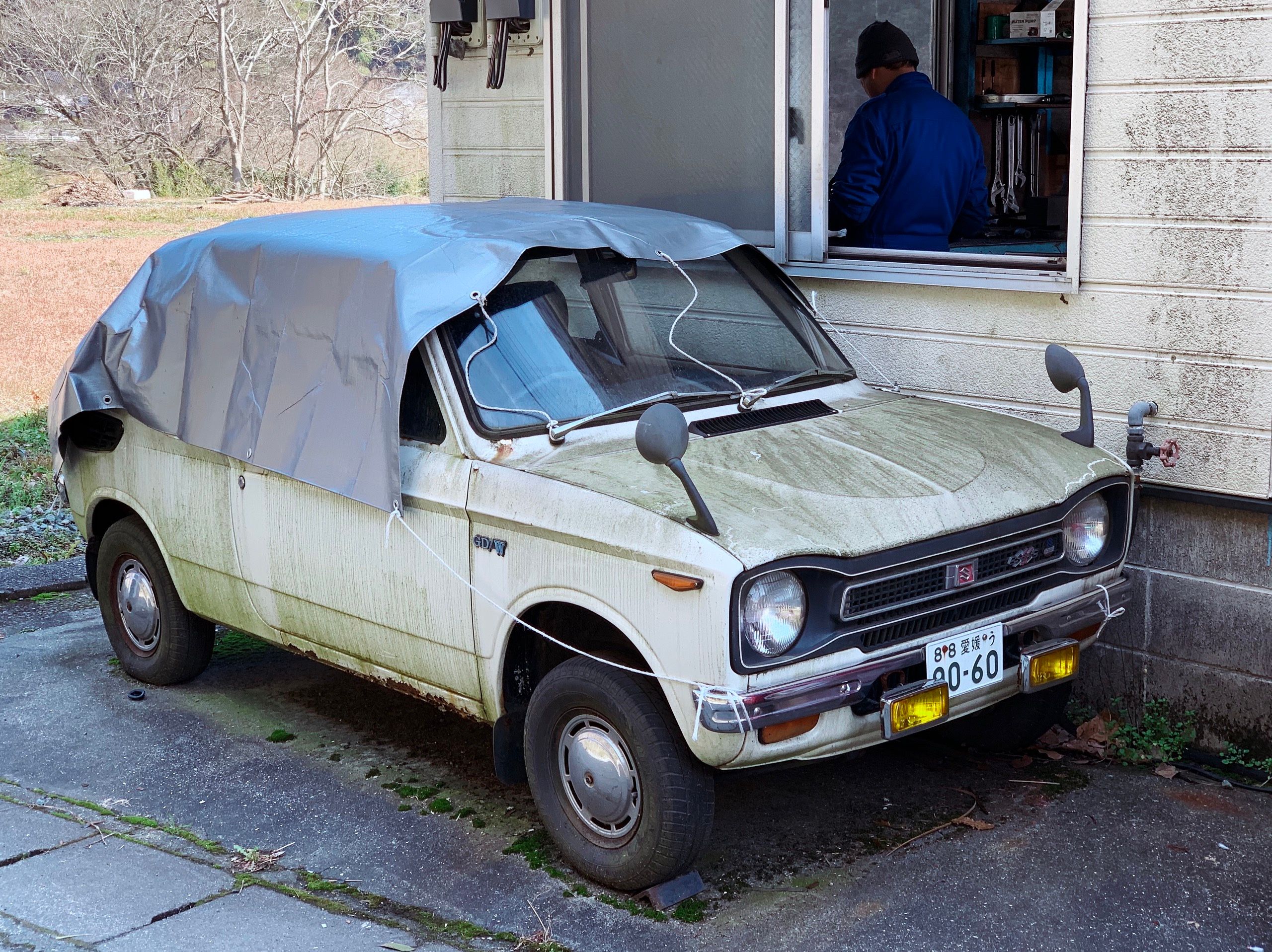 An even smaller Suzuki hatchback, covered with a tarp.