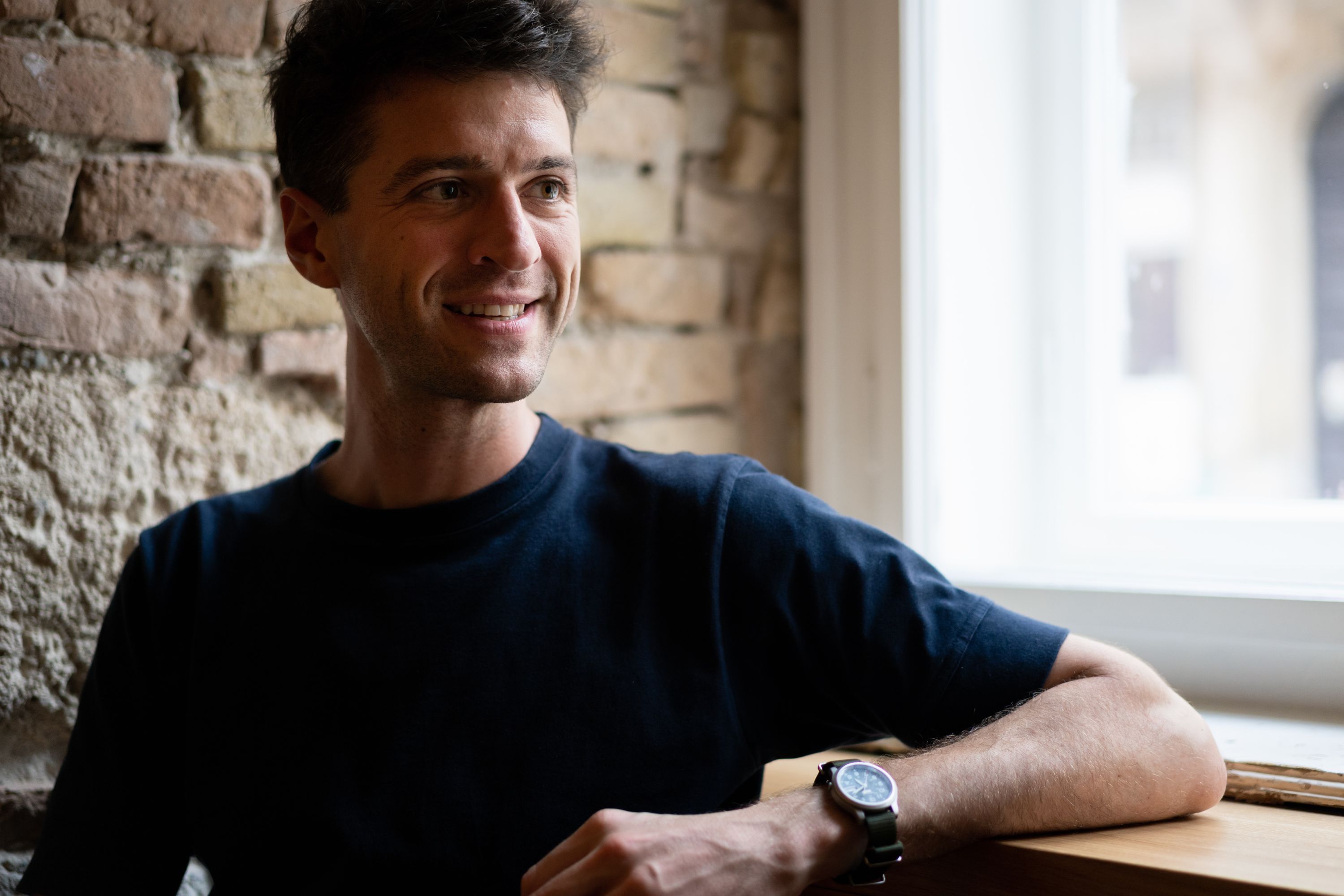 Peter Orosz, a guy with dark hair wearing a navy blue t-shirt, smiling while sitting next to an exposed brick wall. He is positioned near a window with natural light coming in from the right side. He is resting his arm on the windowsill, and he’s wearing a watch with a dark face. The setting has a warm, casual atmosphere with the rustic brick wall contrasting with the bright window light.