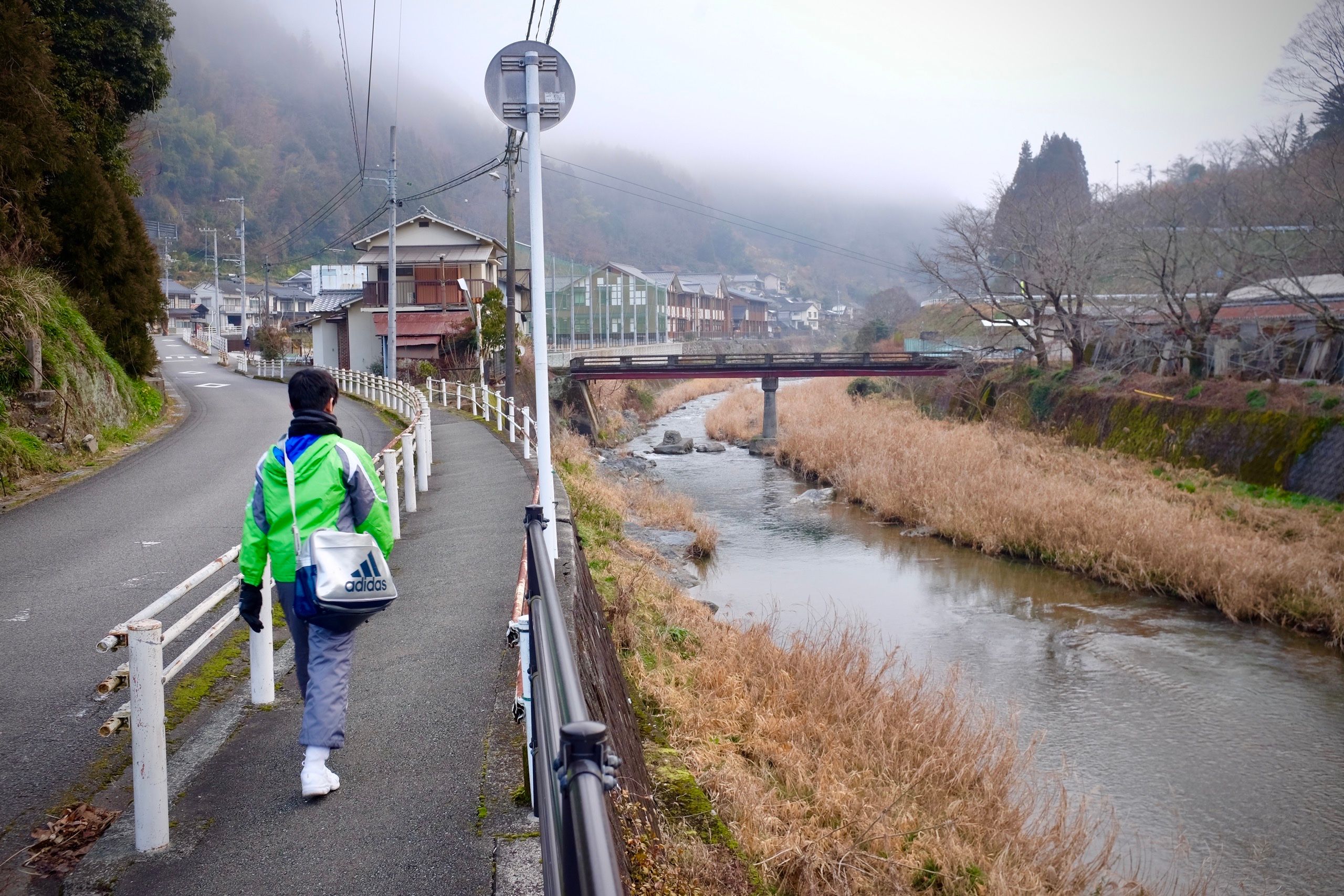 A schoolboy in a green jacket walks down a sidewalk by a stream.