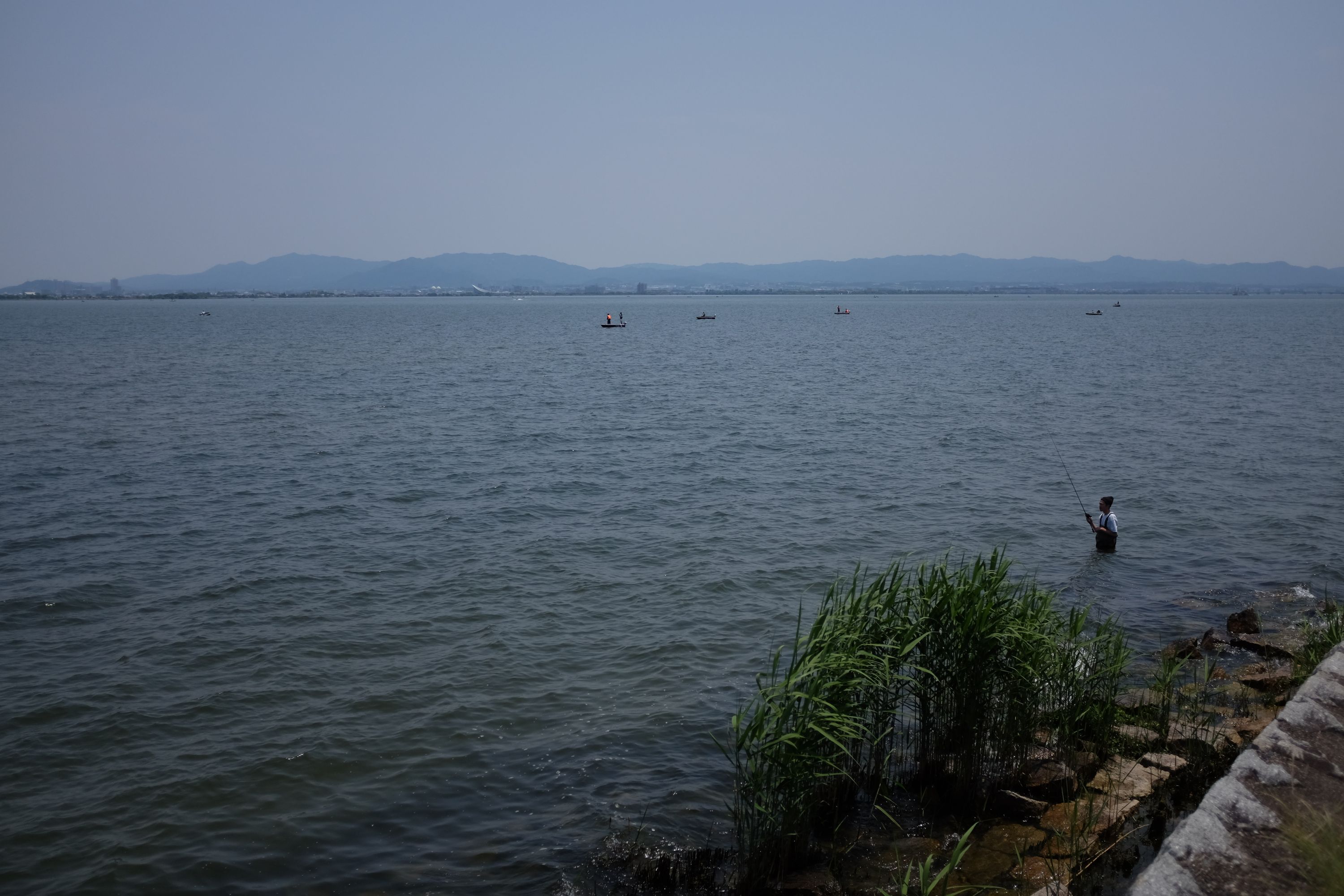 A man in rubber overalls fishing in a large lake.