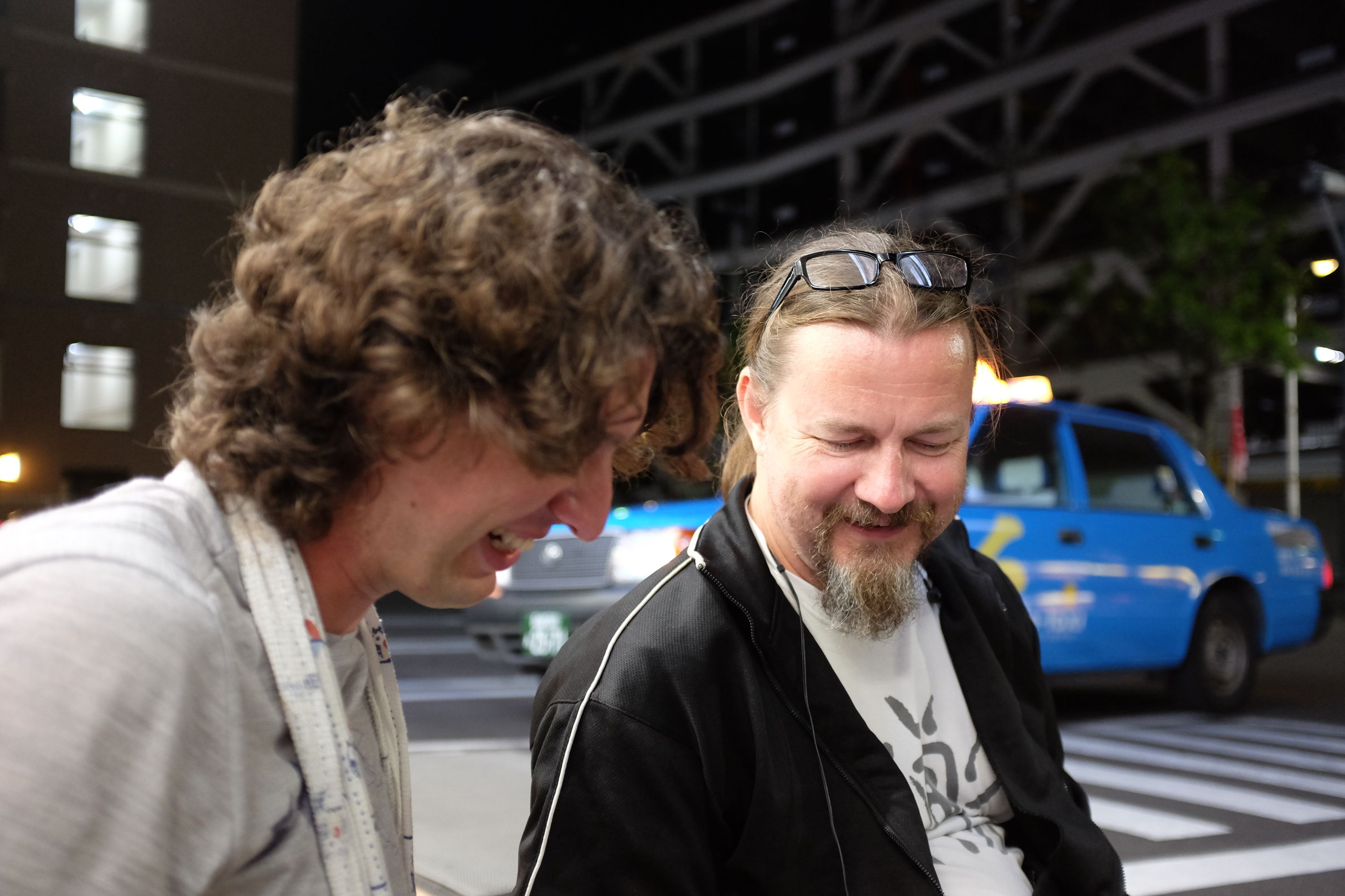 Two men, Gabor and John, laugh in a parking lot.