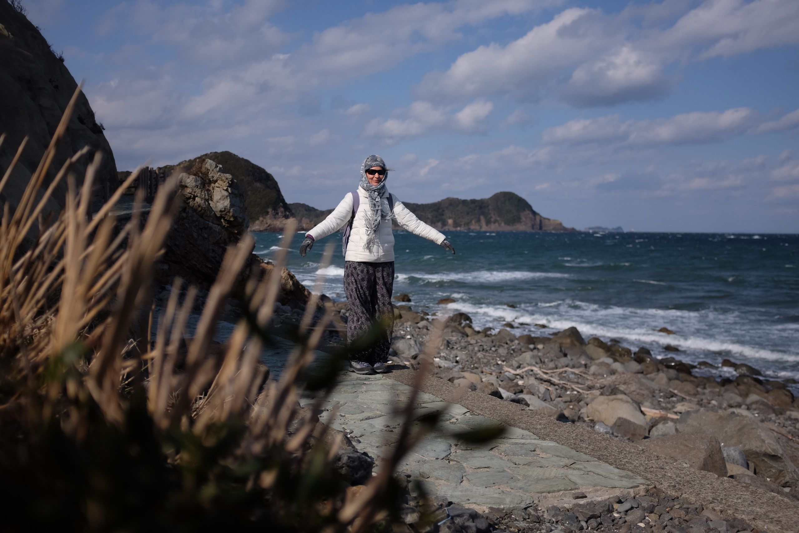 The same woman, Hanga, balances on rocks on a beach, bundled up in many warm clothes.