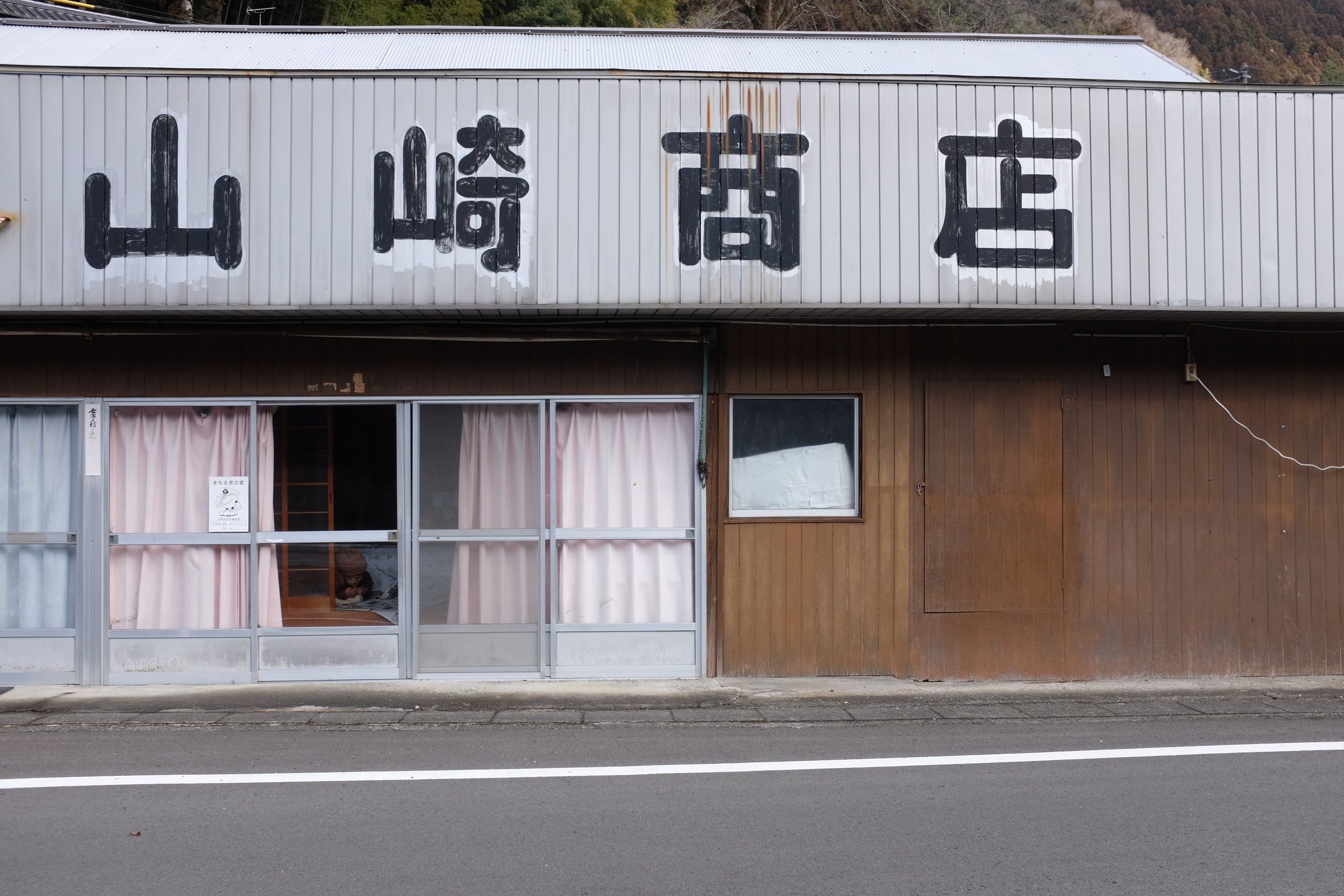 An older woman seen through the open curtain of a building with a sign out front that translates to Yamazaki’s Shop.