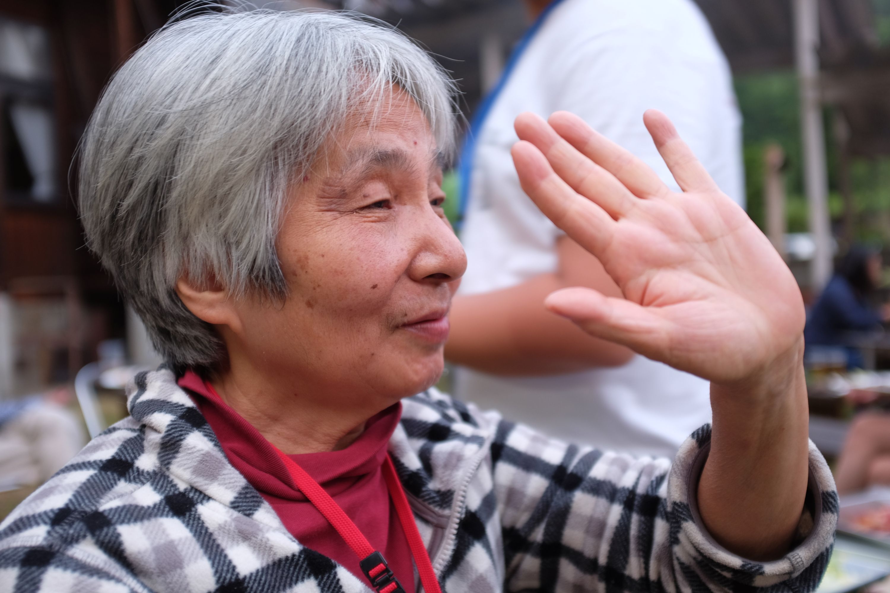 A Japanese woman with short gray hair raises her left hand to wave.