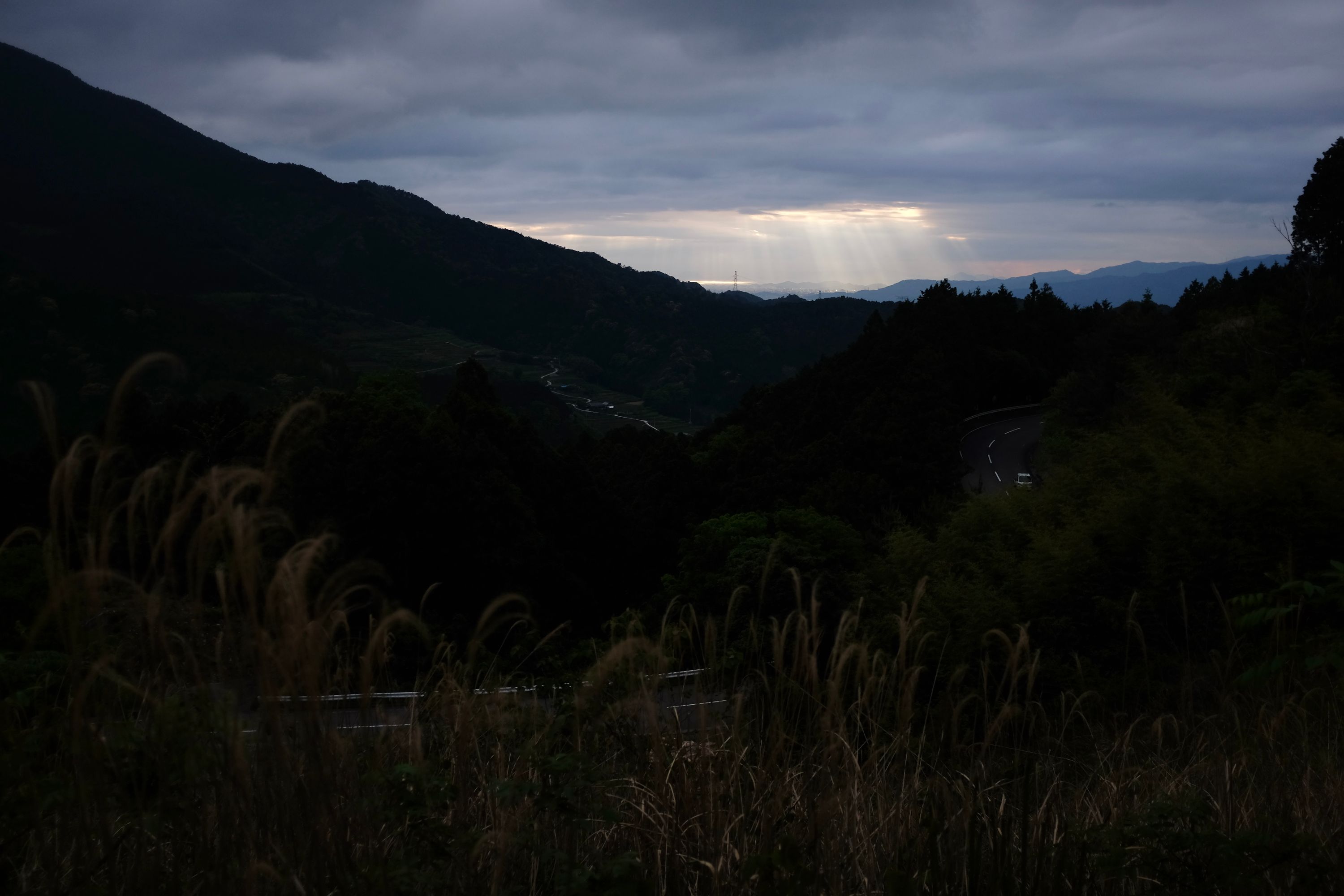 Rays of sunlight pierce the clouds in the distance as seen from a mountain road, outlining a city and the sea beyond.