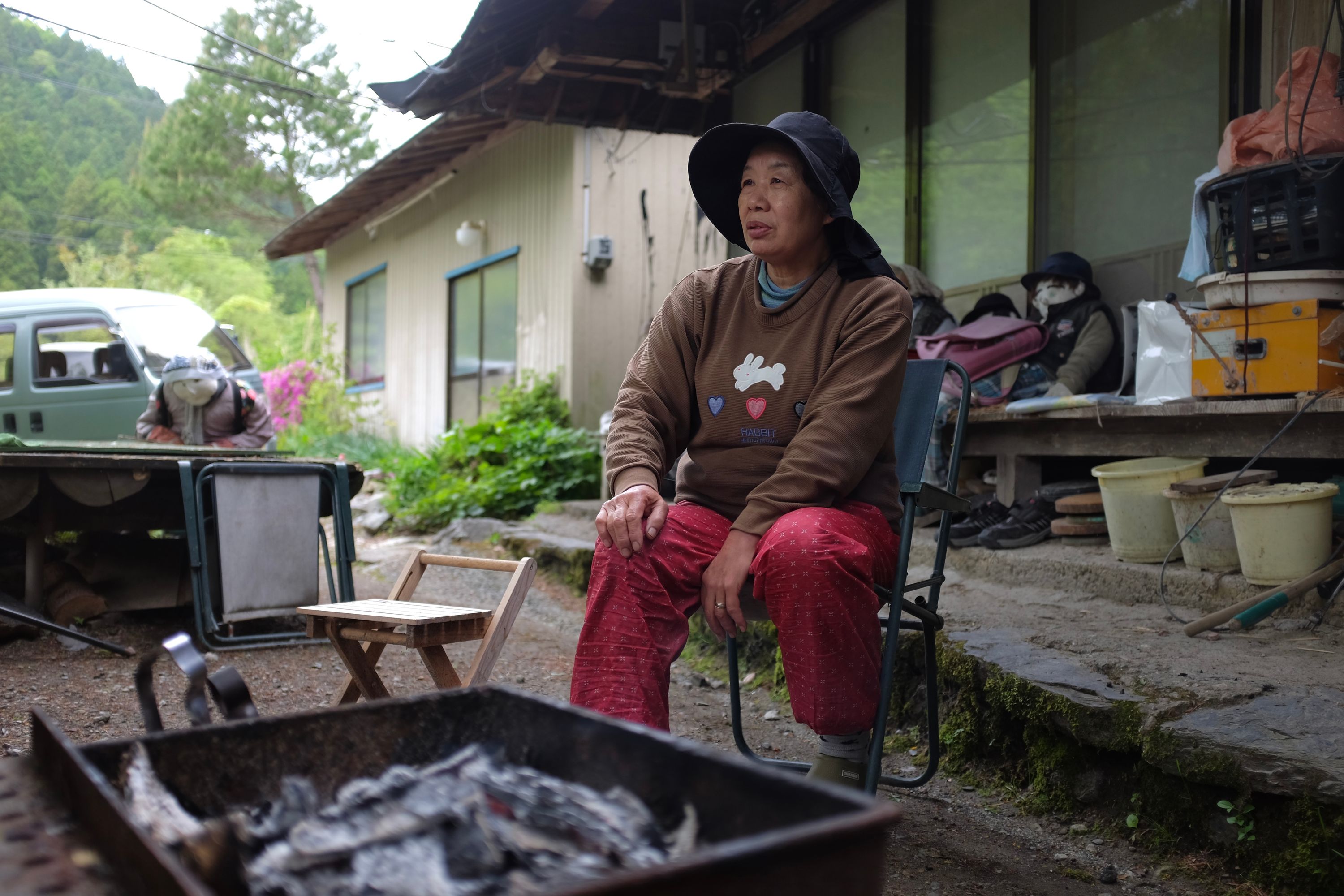 A Japanese woman, Ayano Tsukimi, wearing a wide-brimmed hat and simple country clothes, sits on a chair in front of her house, with several of her dools visible behind her.