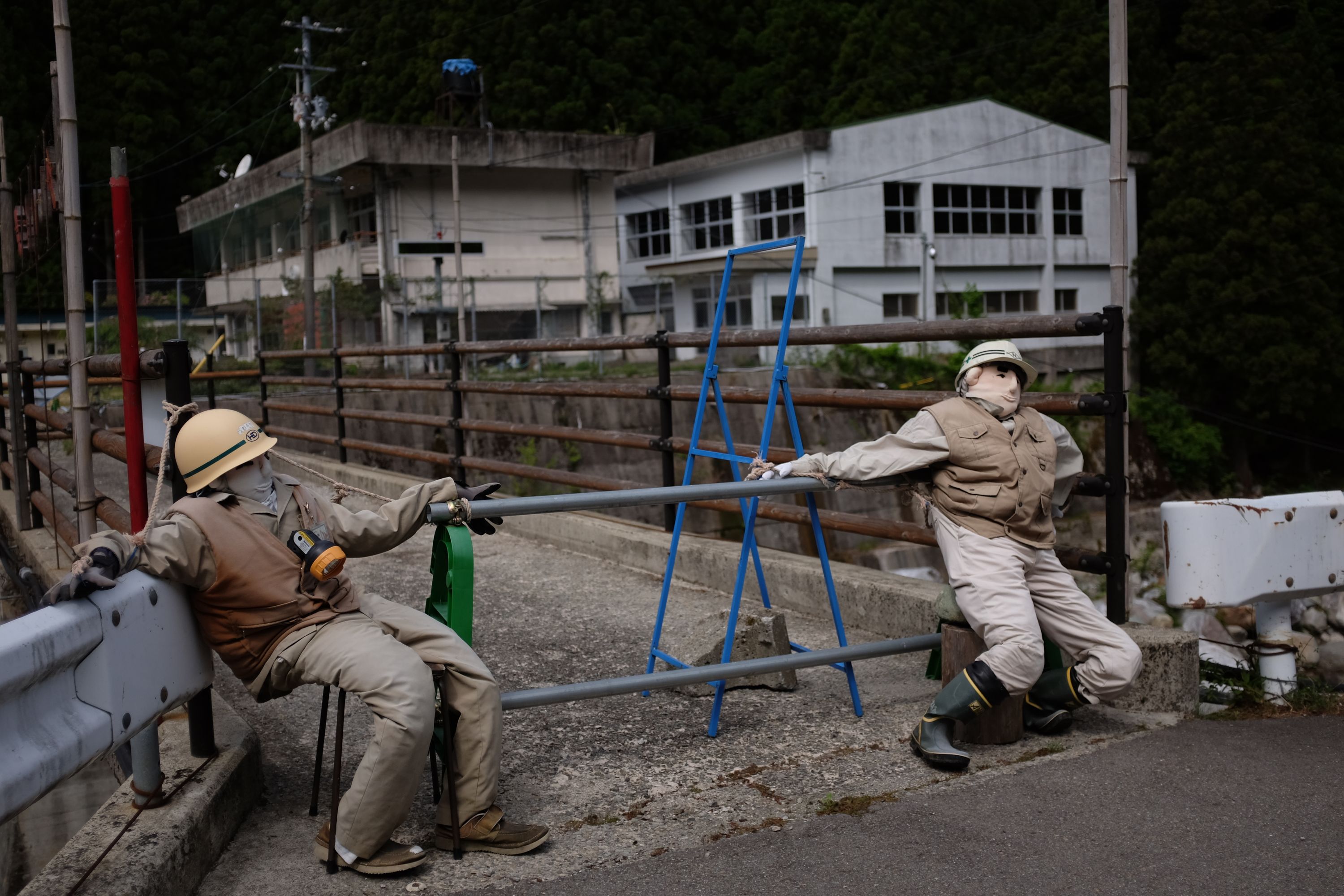 Two dolls wearing hard hats guard a closed bridge leading to an abandoned schoolhouse.