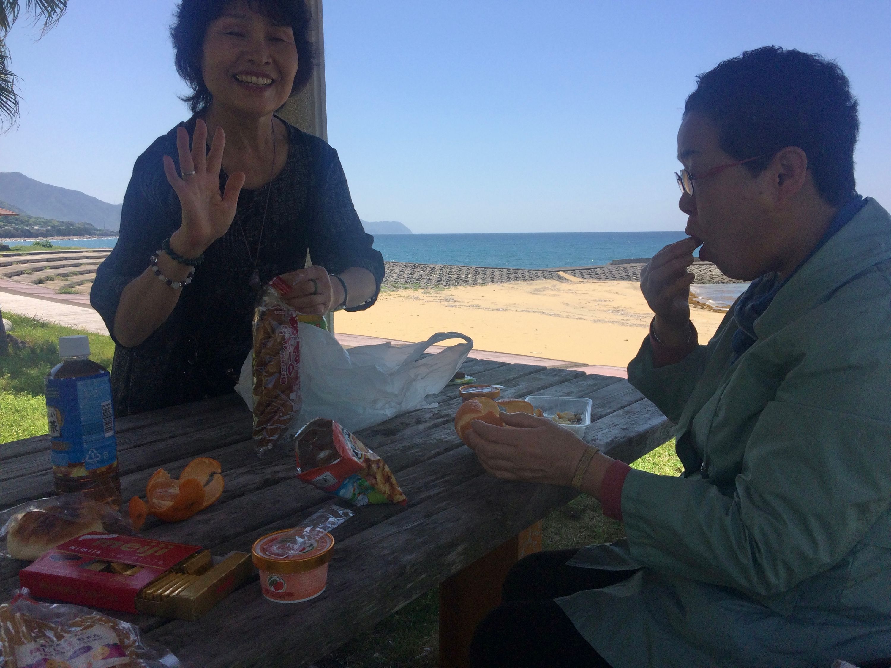 Two women having lunch at a picnic table, one of them waves at the camera.