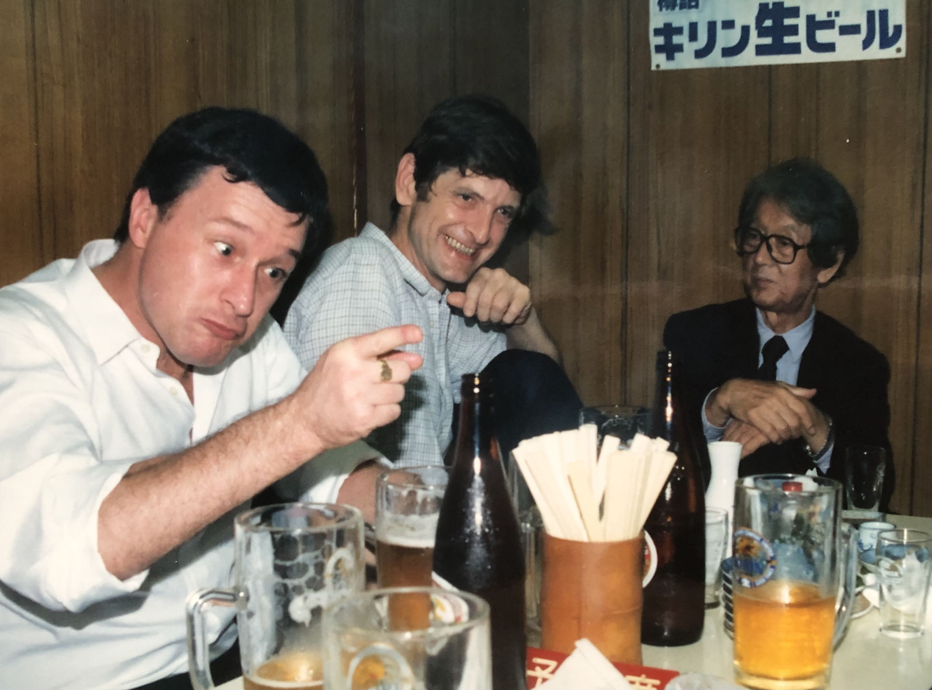 A color photograph showing three men seated at a table in a Japanese restaurant or bar. On the left is Alan Booth, an Englishman with dark hair wearing a white polo shirt, pointing at the camera with a slightly mischievous expression. In the center is Timothy Harris, another Englishman with dark hair wearing a light blue checkered shirt, smiling broadly. On the right is an older Japanese man wearing glasses, a dark suit and tie, sitting more formally. The table has multiple beer glasses and a dark bottle visible. Behind them is a wood-paneled wall with Japanese text on a white sign that reads “キリン生ビール” (Kirin Draft Beer). The scene captures a casual drinking session in 1980s Tokyo.