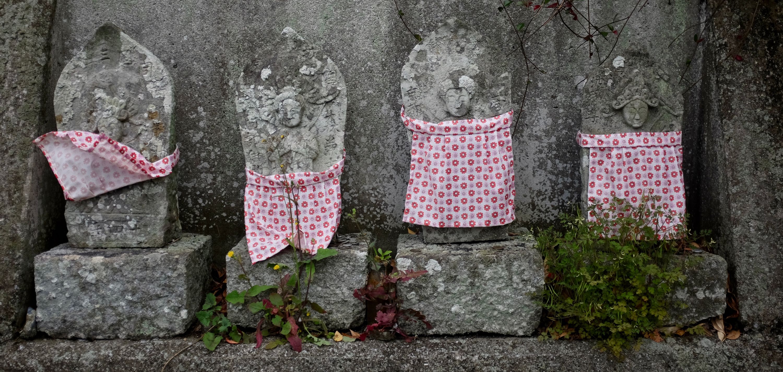 Four small roadside Buddhist statues wearing aprons, with the apron of one flipped up by the wind.