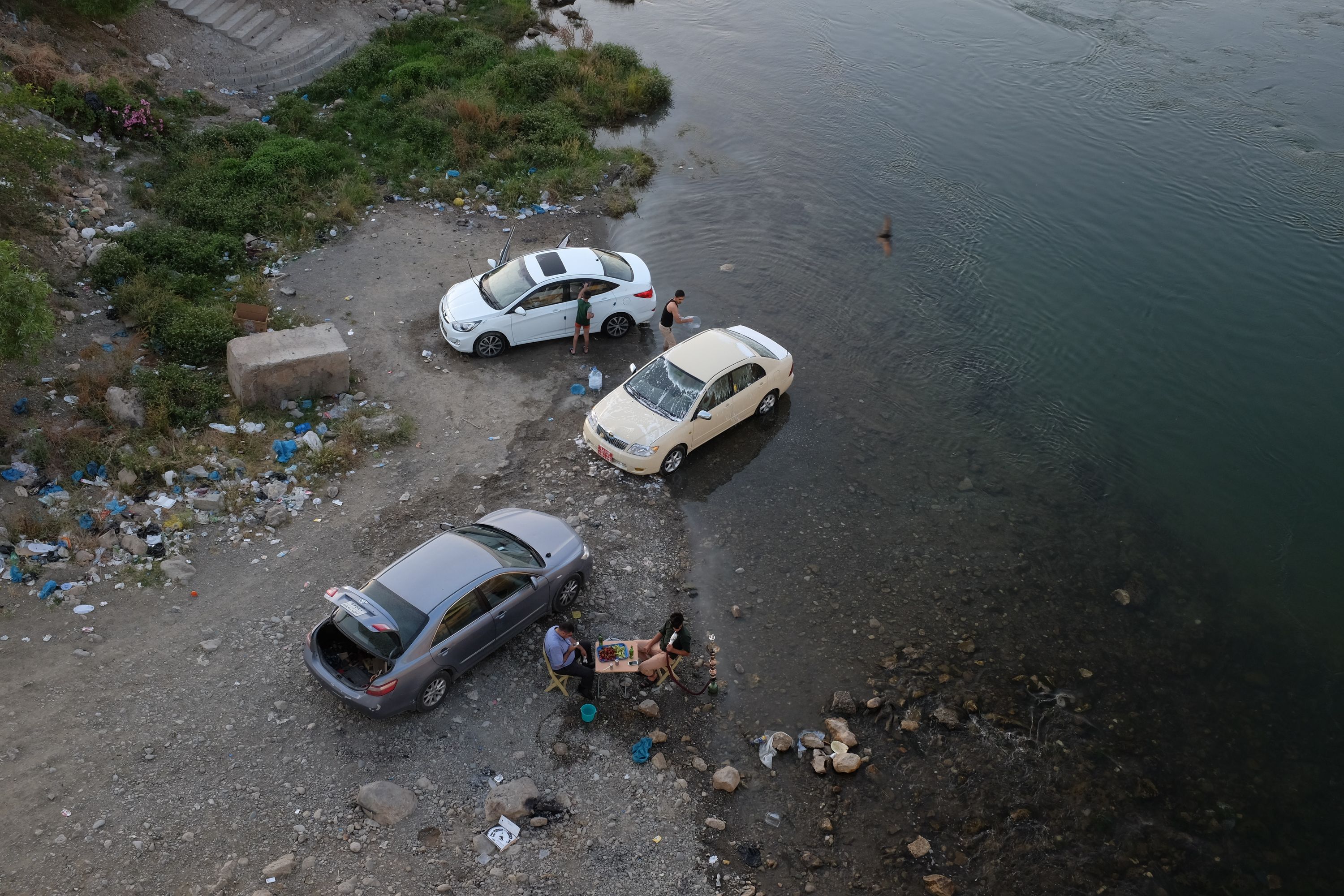 Looking down from a bridge at people washing their cars and having picnics by a river.