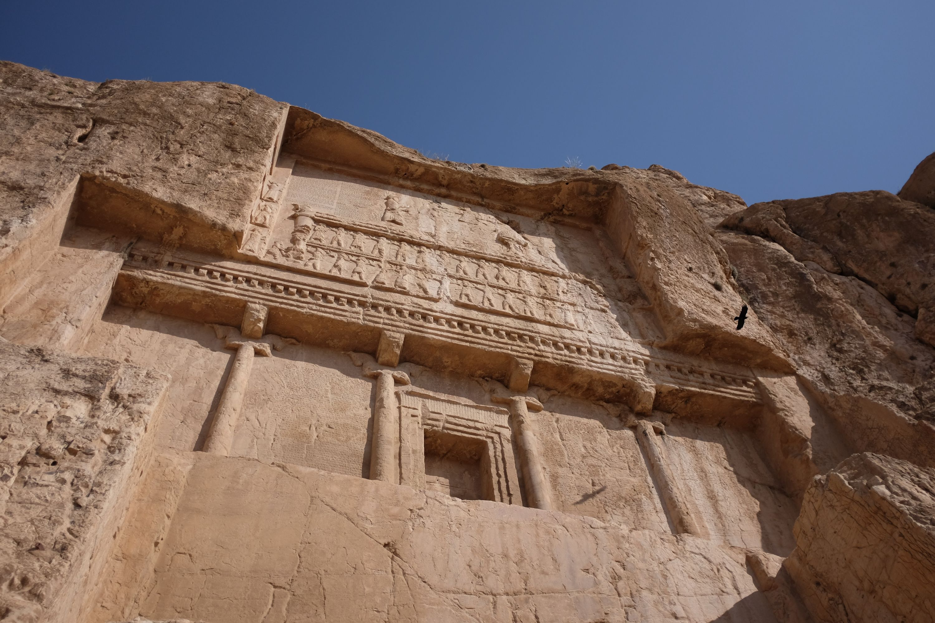 Looking up at the entrance of a large tomb carved into a vertical rock wall.