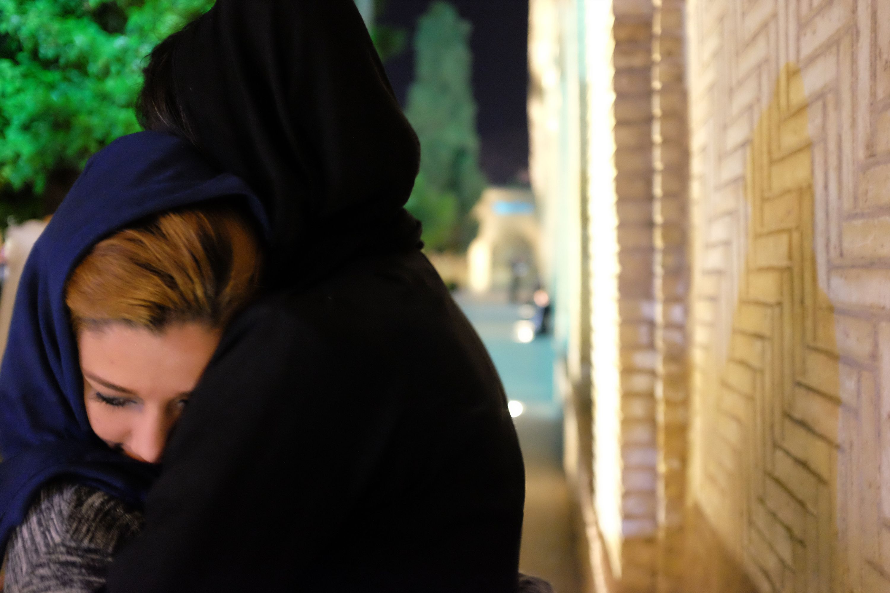 A woman in a dark headscarf, only her silhouette visible, hugs a woman whose hair peeks out from under her headscarf, in front of a yellow brick wall.