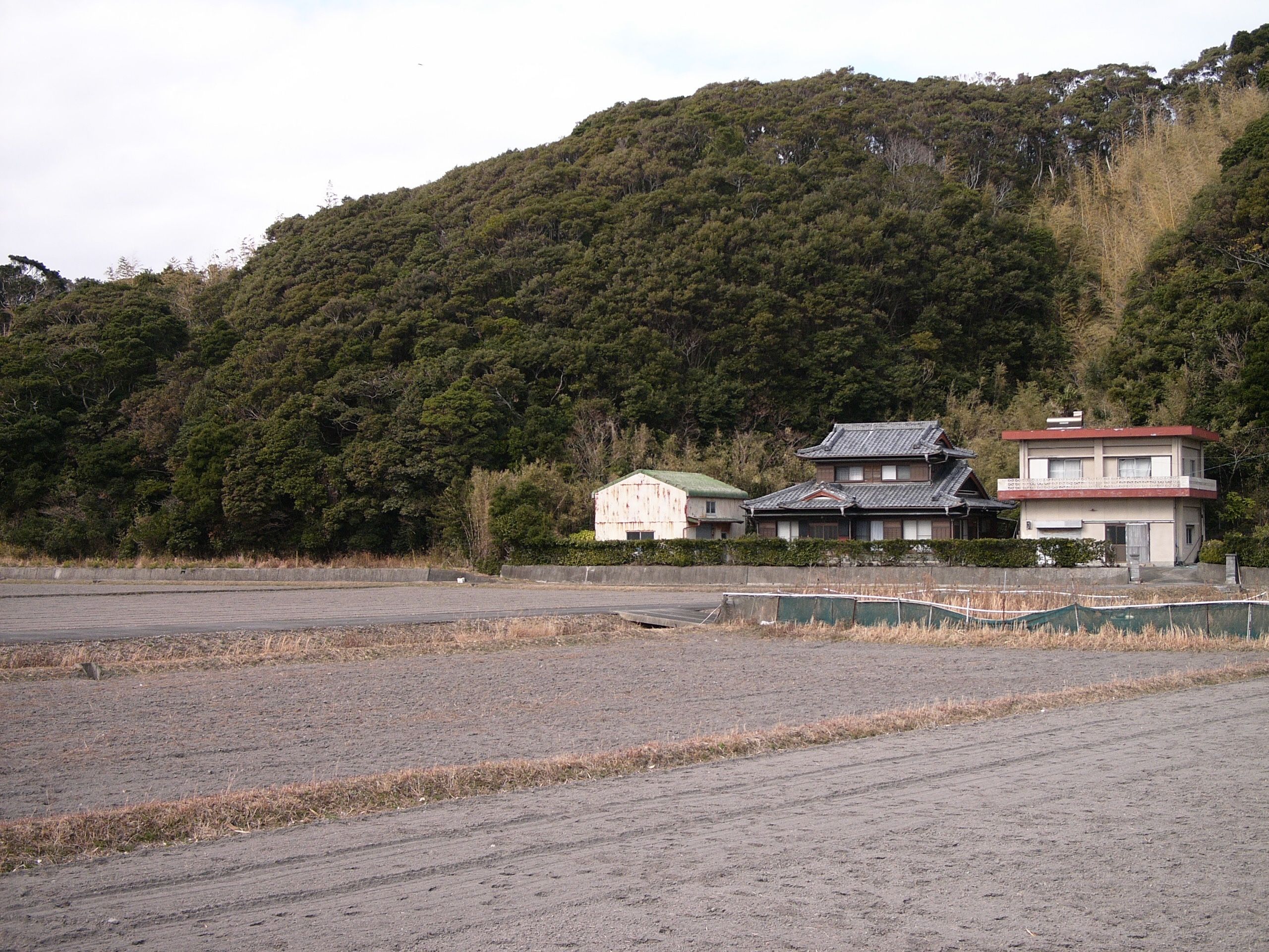 Three farmhouses stand against a hillside in front of fallow fields.