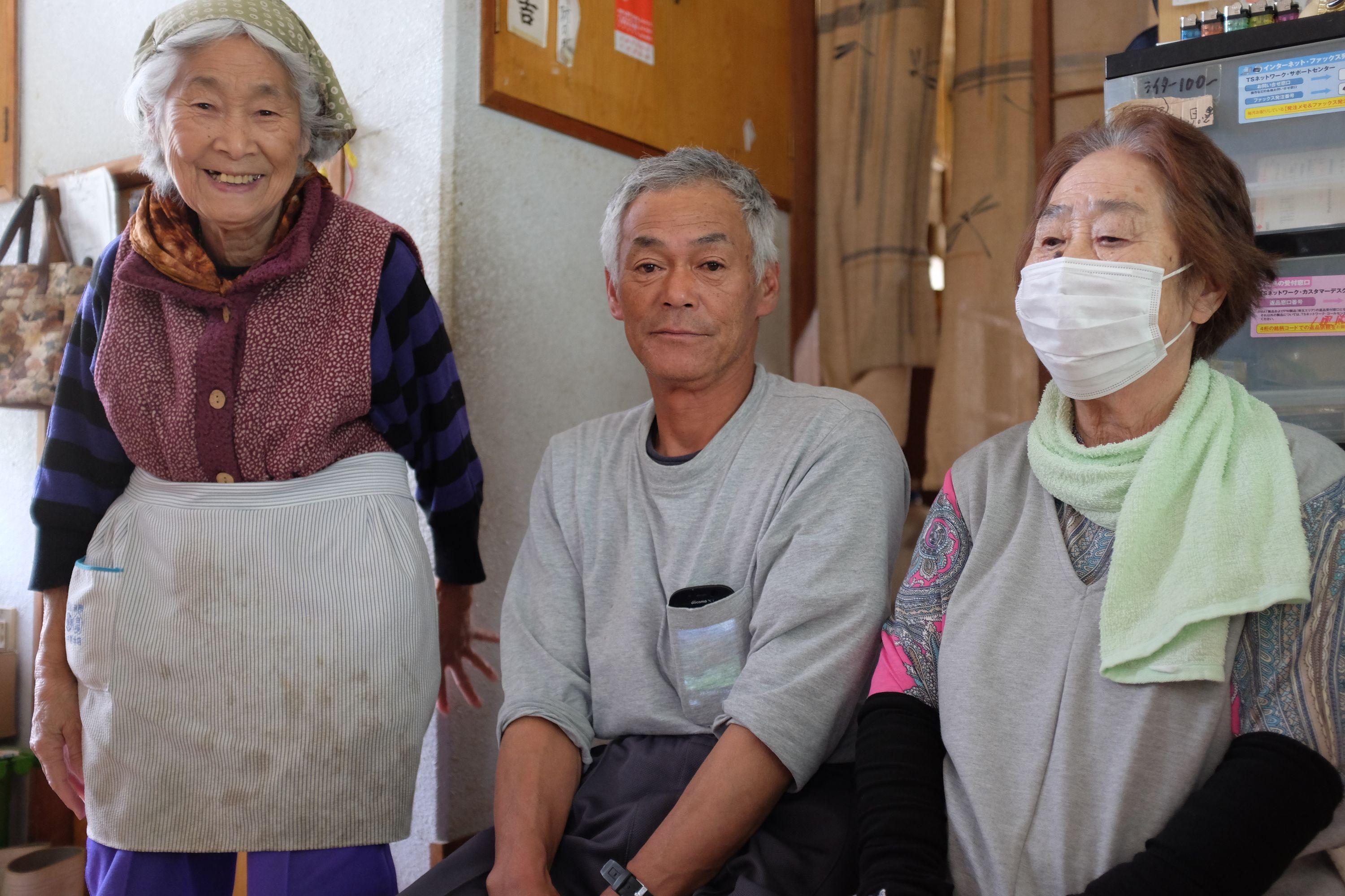 A smiling elderly shopkeeper poses for the camera with her husband and one of their customers.