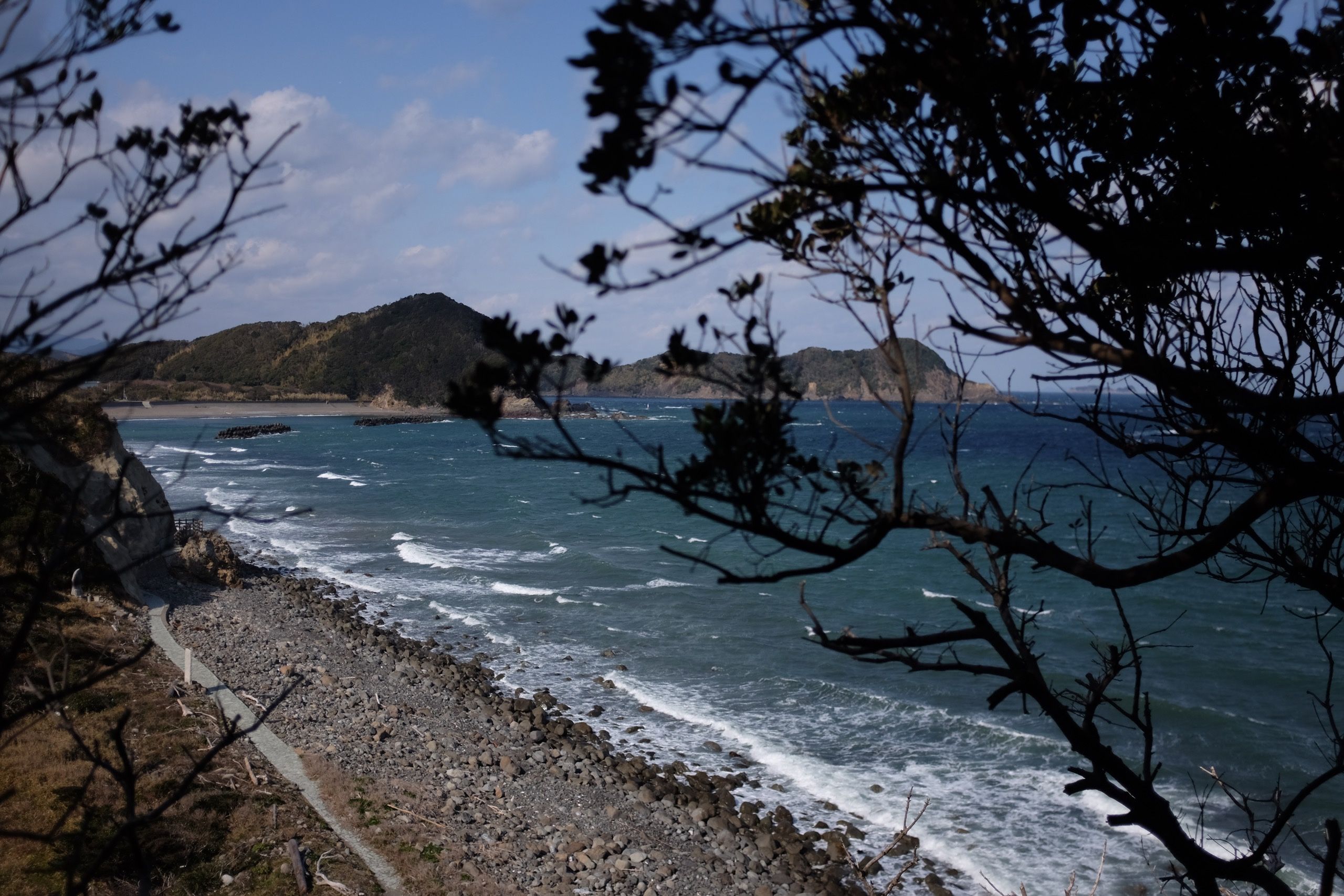 A rocky beach as seen from between small trees, with more forested hills on the horizon.