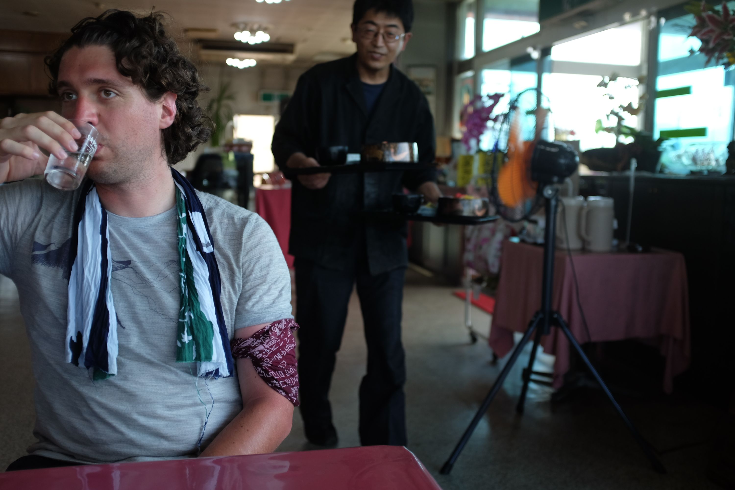 Gabor drinks a glass of water in a restaurant as a waiter approaches with two trays of food.