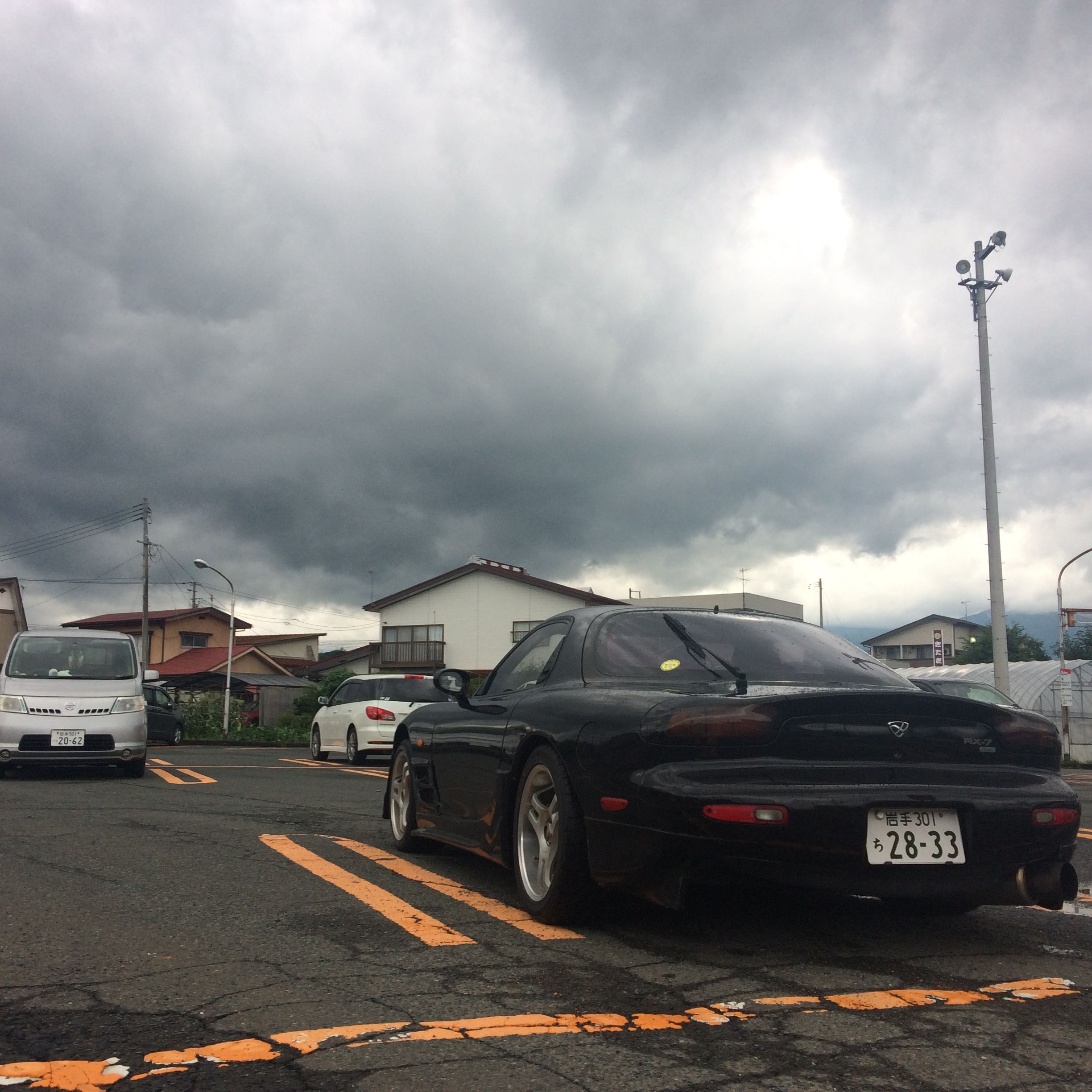 A black Mazda RX-7 sports car in a parking lot under dark grey clouds.