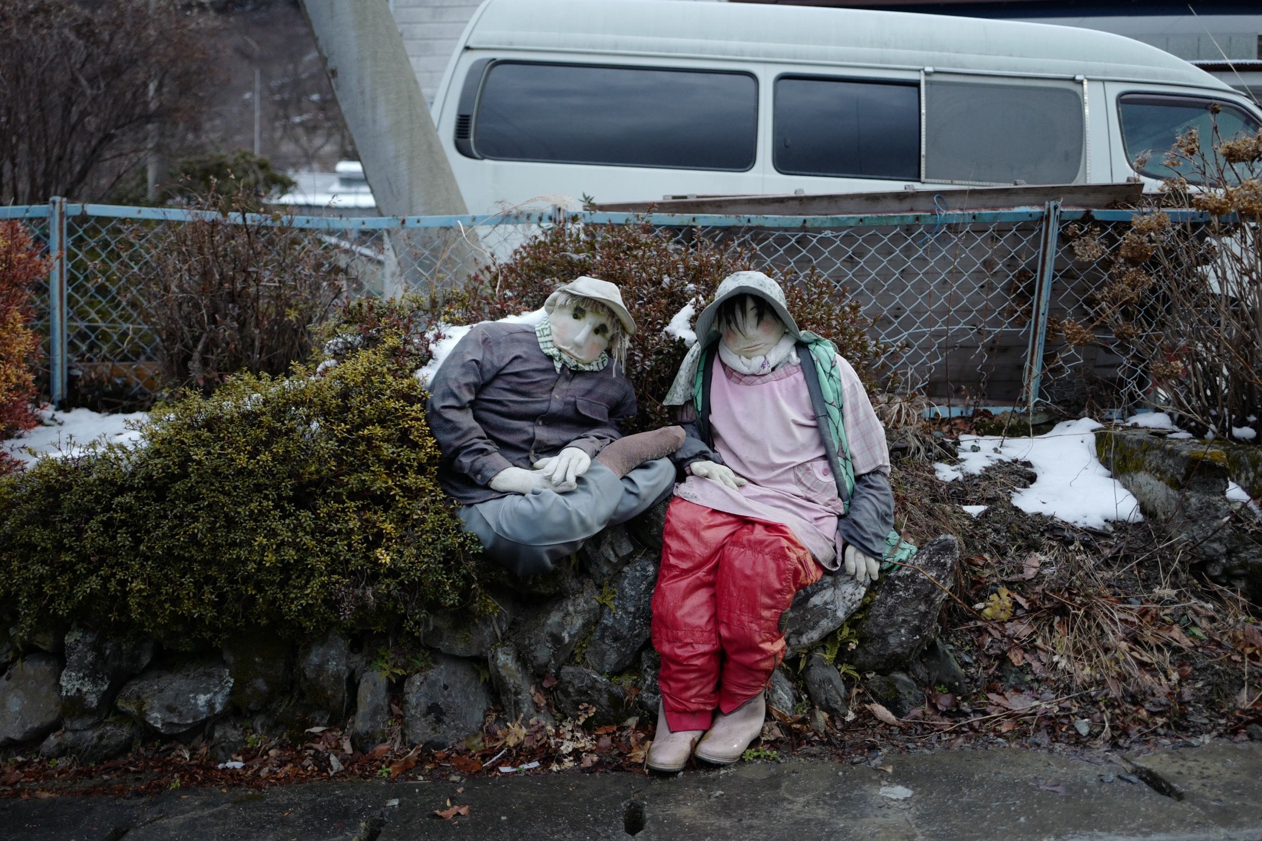 Two similar dolls sit on a low rock wall, they are partially covered in snow.