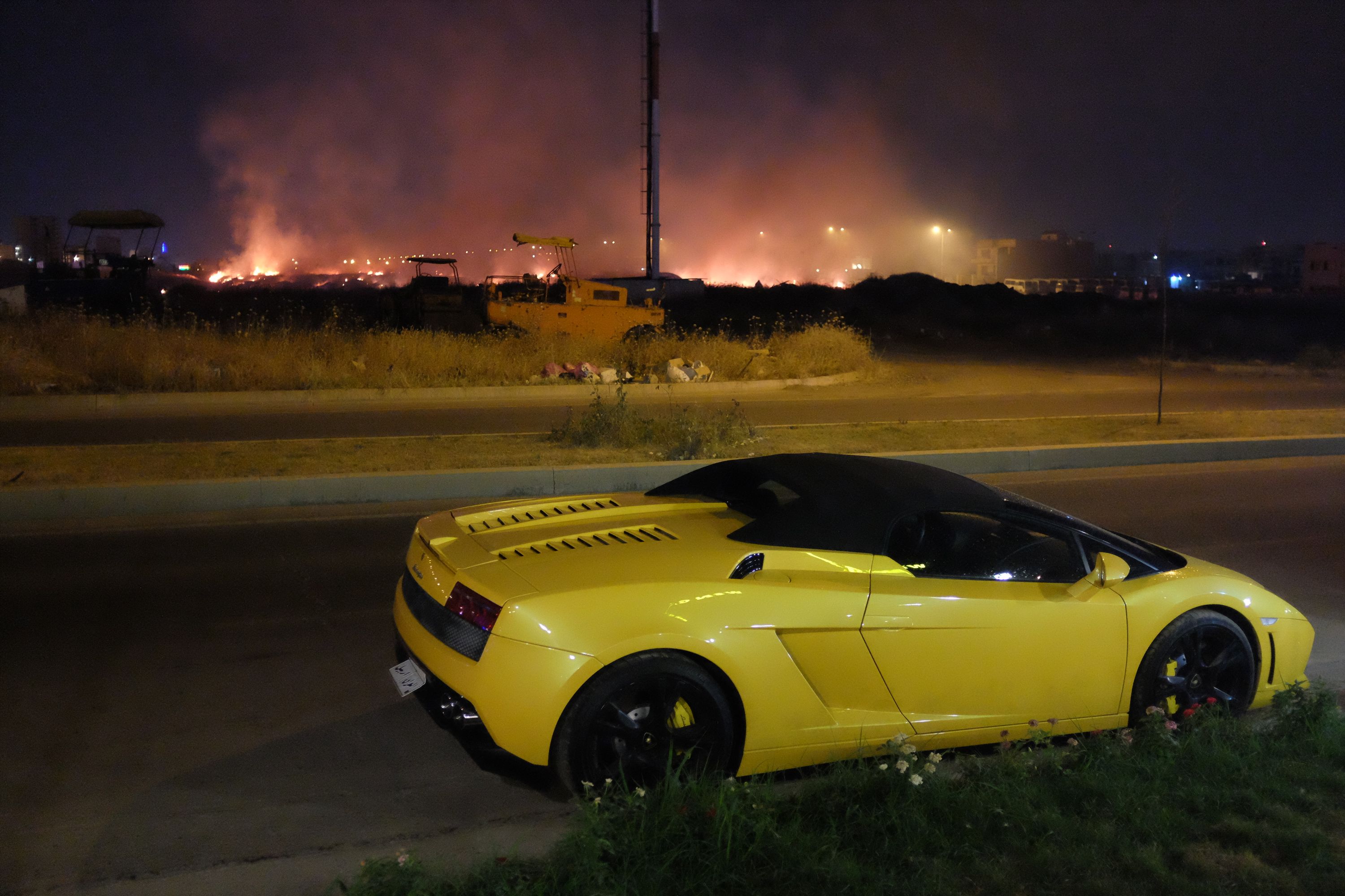 A yellow Lamborghini Gallardo parked across the road from what looks like an enormous trash fire which lights up the night sky.