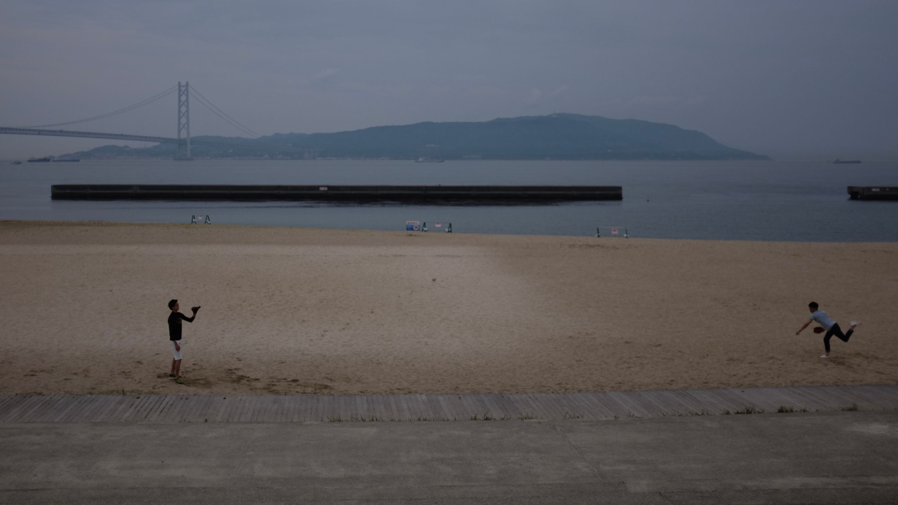 Two boys play baseball on a beach with the Akashi Kaikyō Bridge in the background.