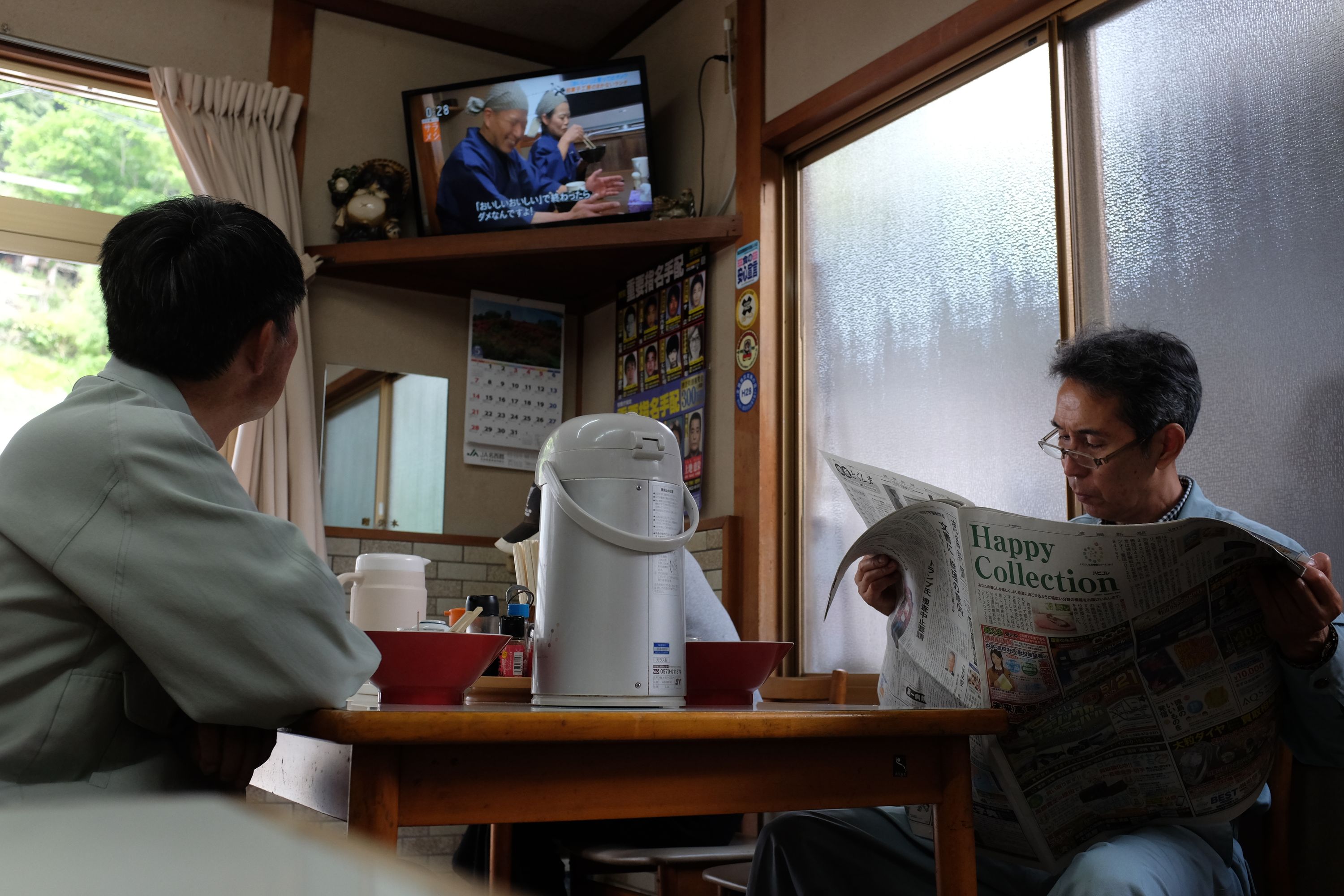 Two men sit at a restaurant table, one watching television, the other reading a newspaper.