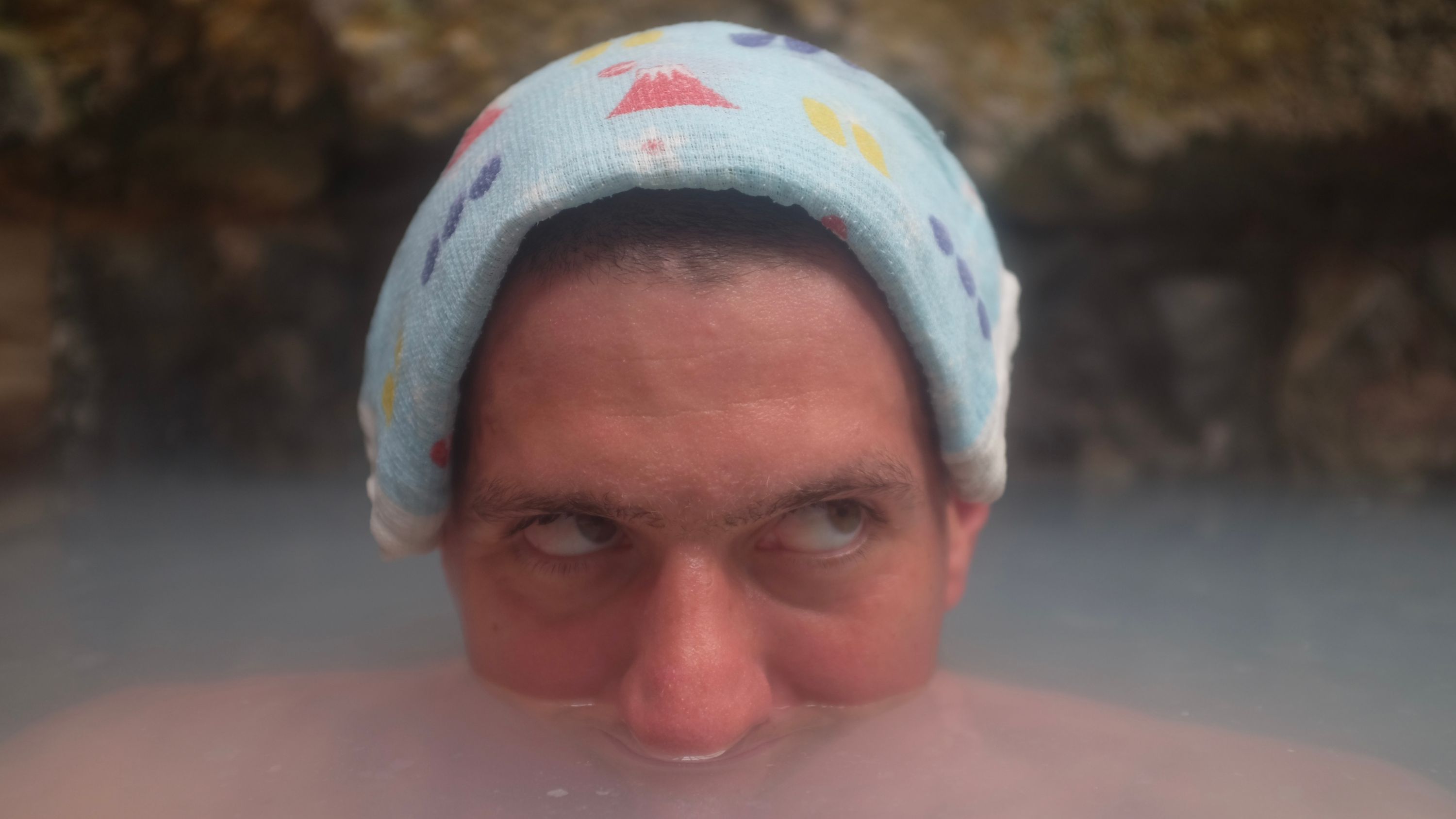 The author in the tub of a hot spring with cloudy blue-white water, submerged to his nose, with a small Japanese towel on his head.