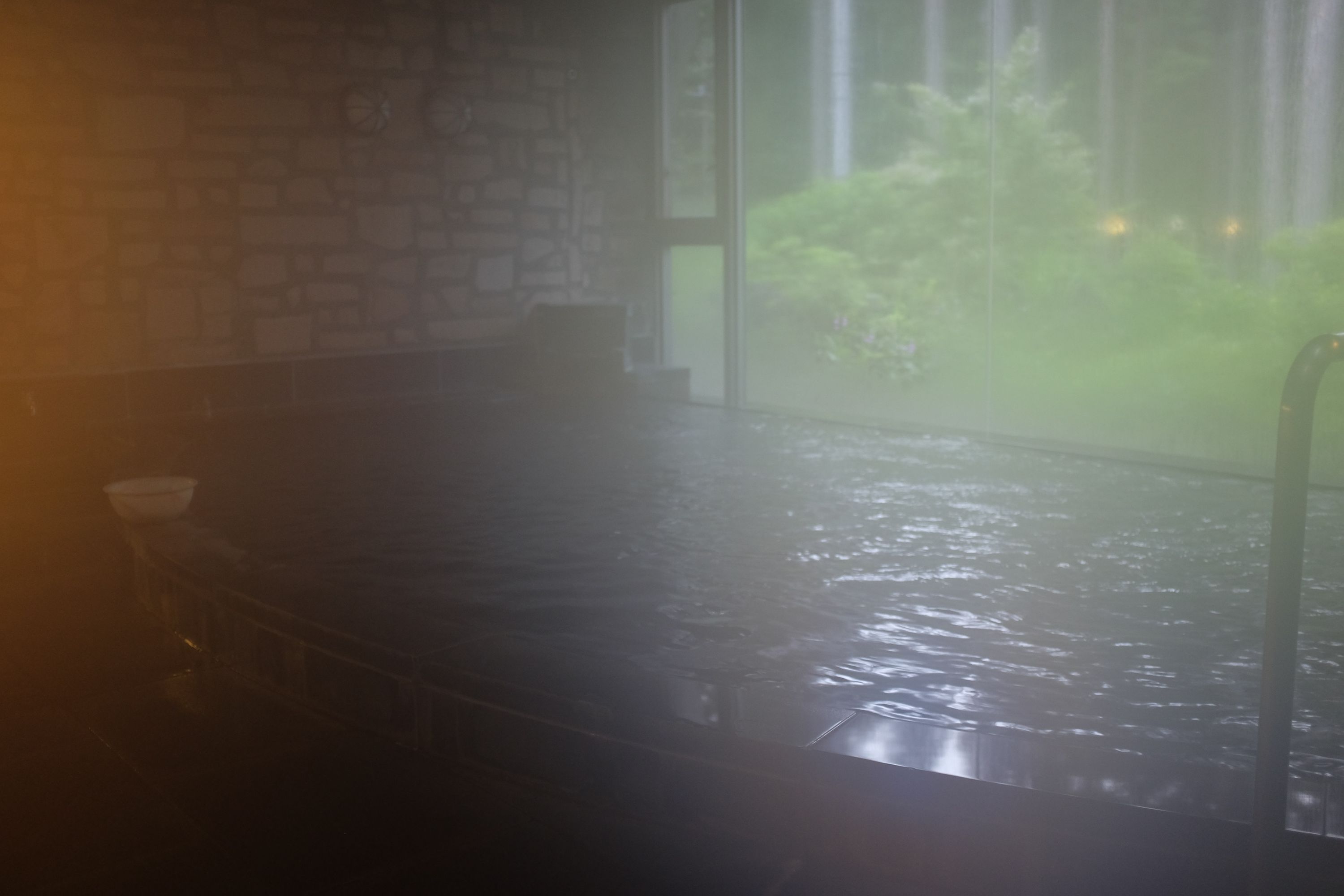 The bathtub of a bathhouse is separated from the forest by a large window, but you can barely see anything from the steam.