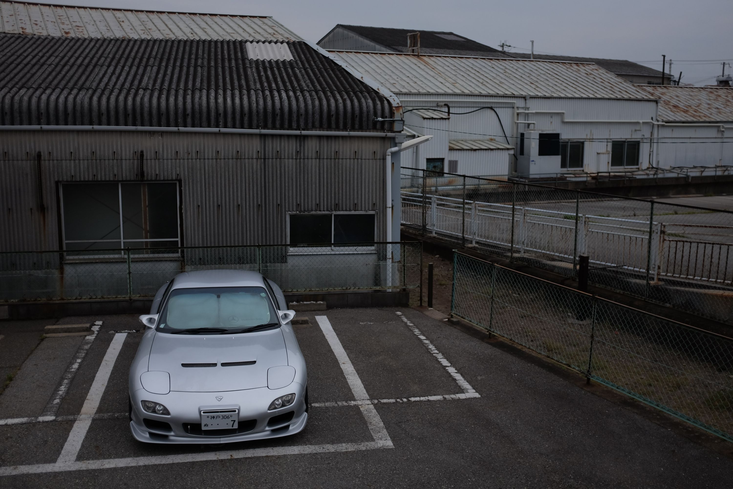 A silver Maxda RX–7 sports car parked in front of gray industrial buildings.