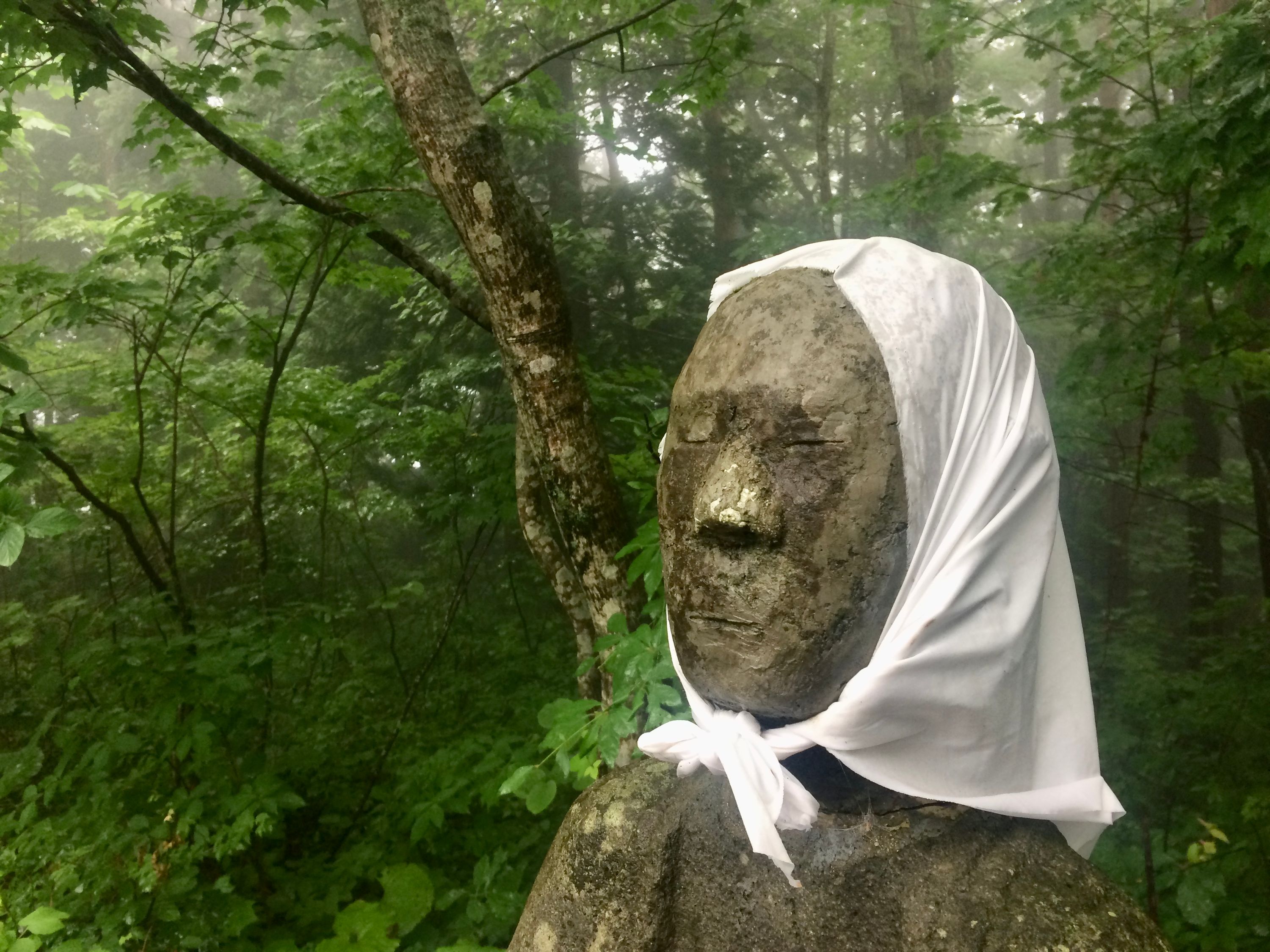 A stone statue of a roadside deity in the forest wears a white headscarf .
