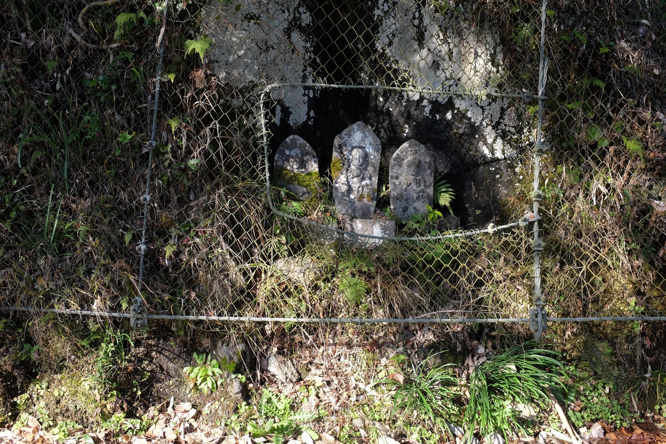Three roadside deities peek through a window cut for them in a chain-link fence protecting against rockfalls.