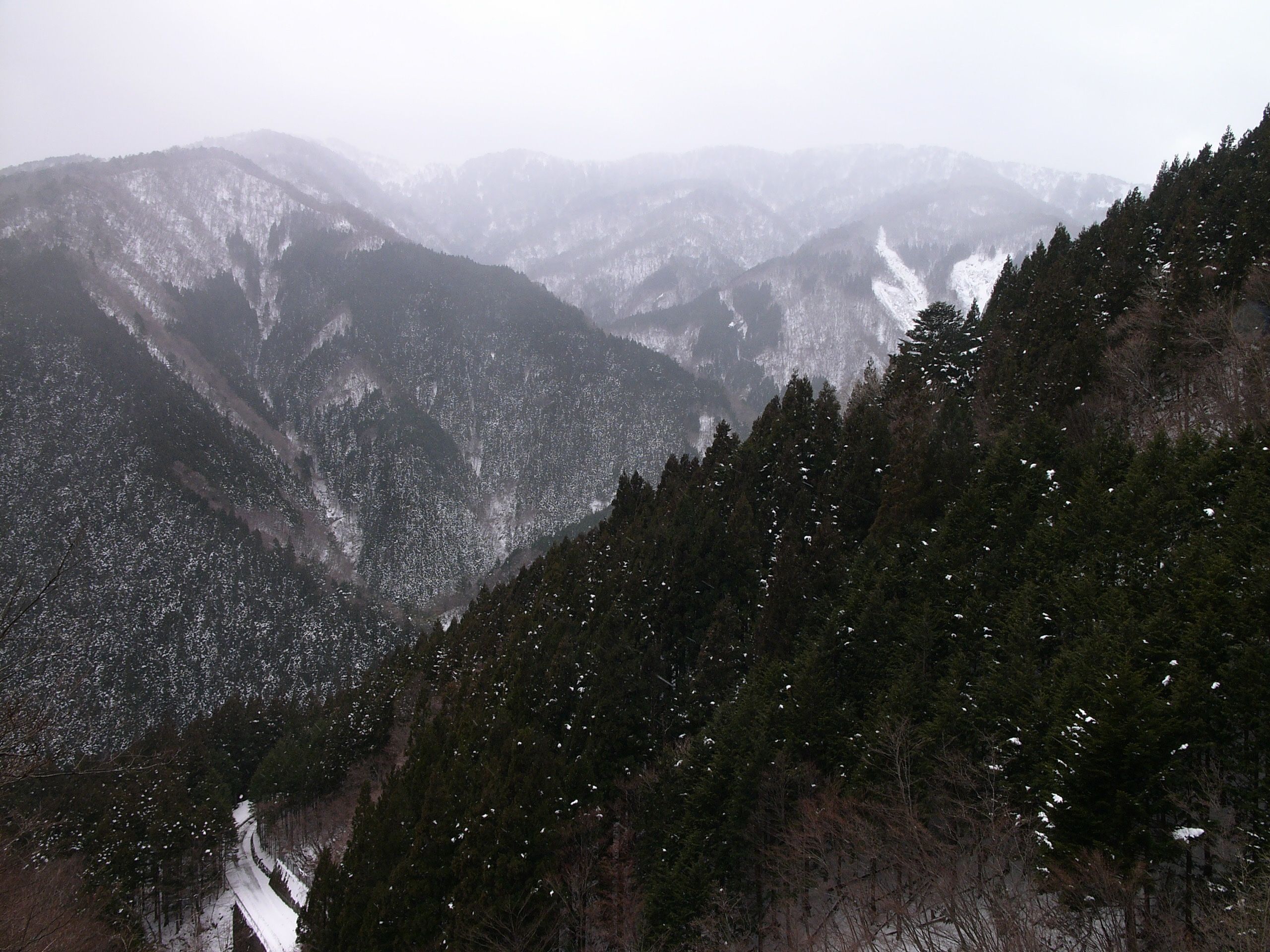 A mountain valley in the winter with a very similar composition: green-black forests, snow clouds in the sky, the only road covered in snow.