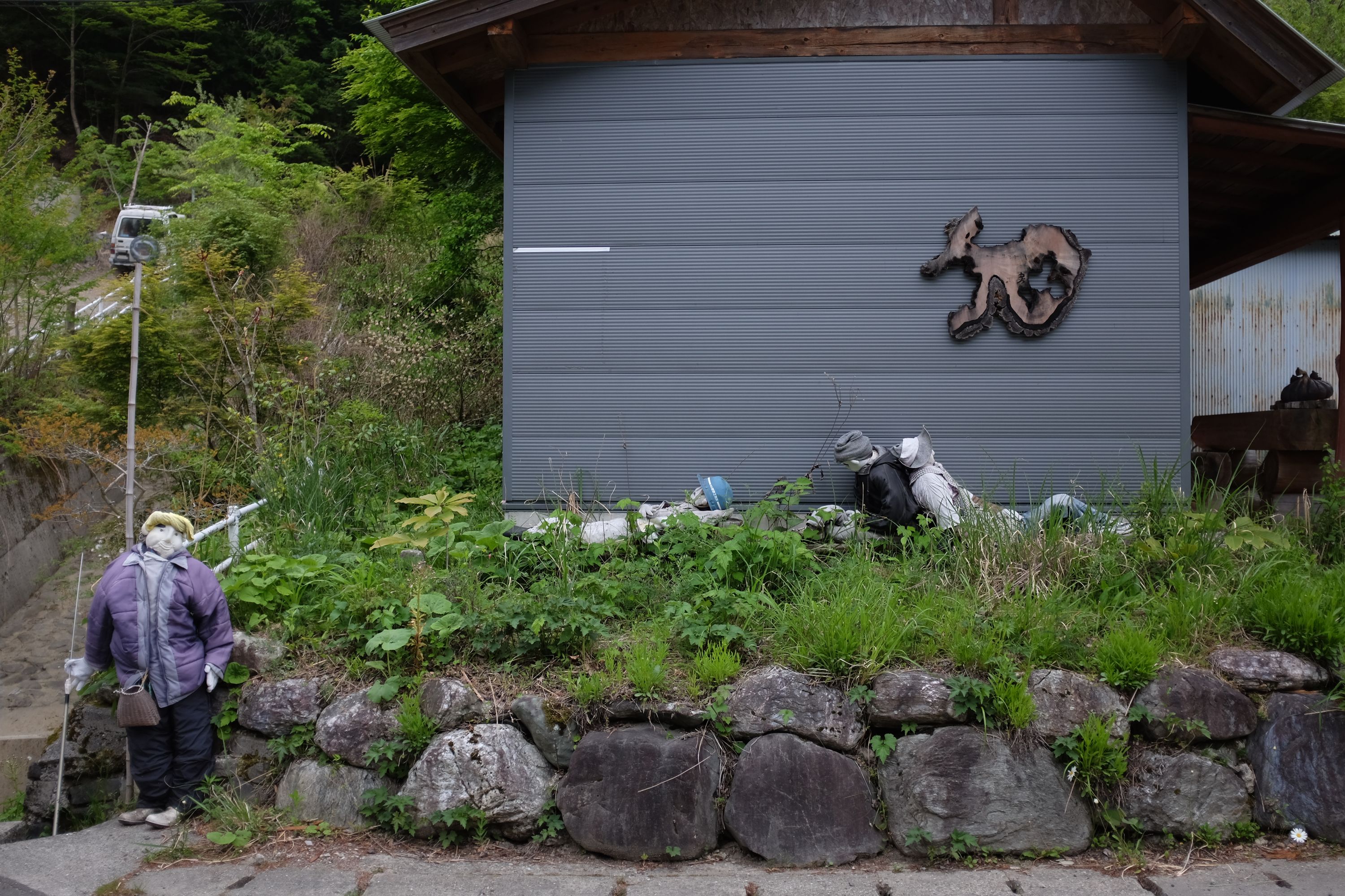 Three dolls lie on the ground in front of a house, while a fourth loiters by the road.