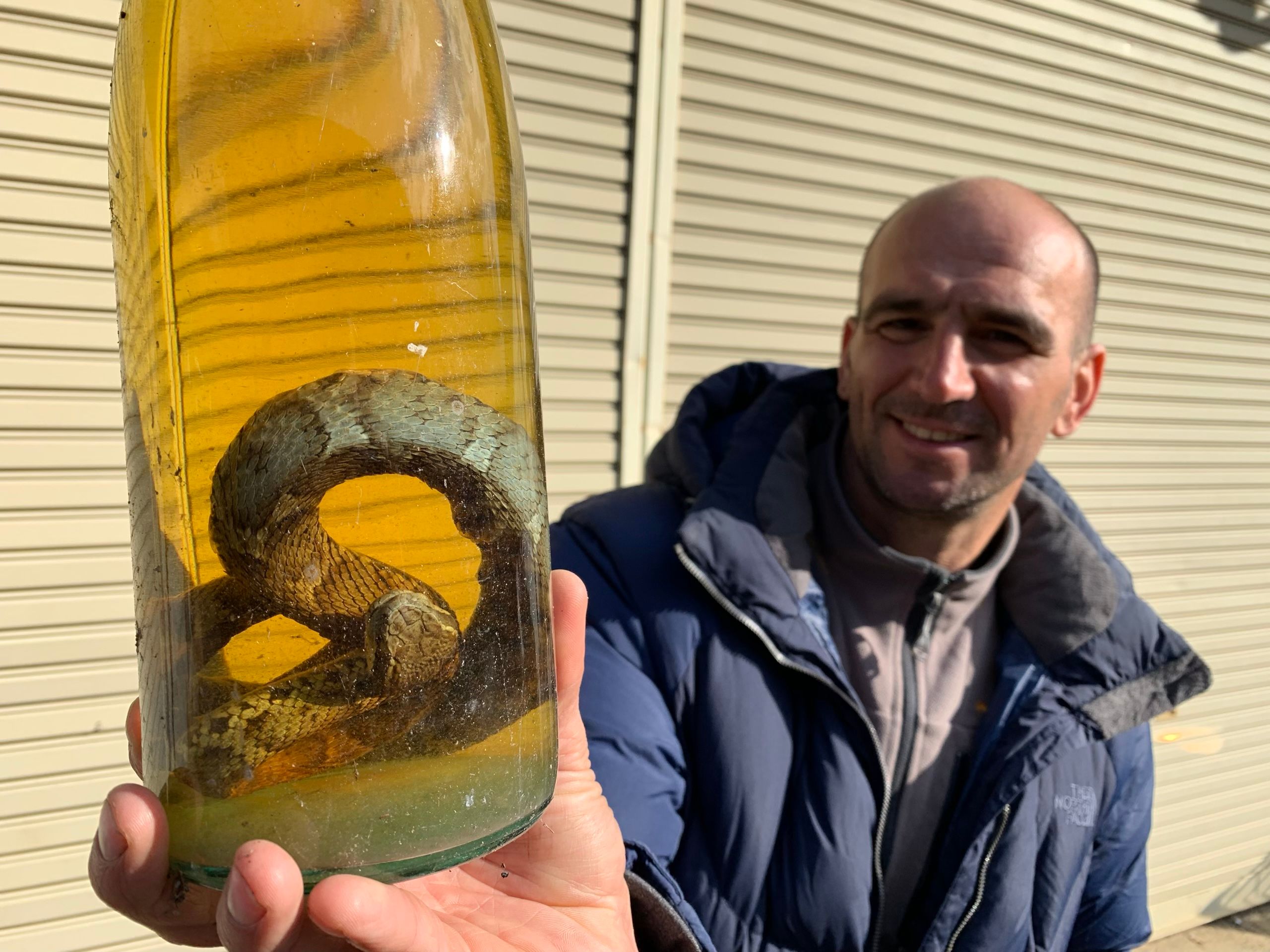 A man in a navy blue parka, Gyökös Lajos, holds up a bottle of yellow liquid with a snake coiled inside.