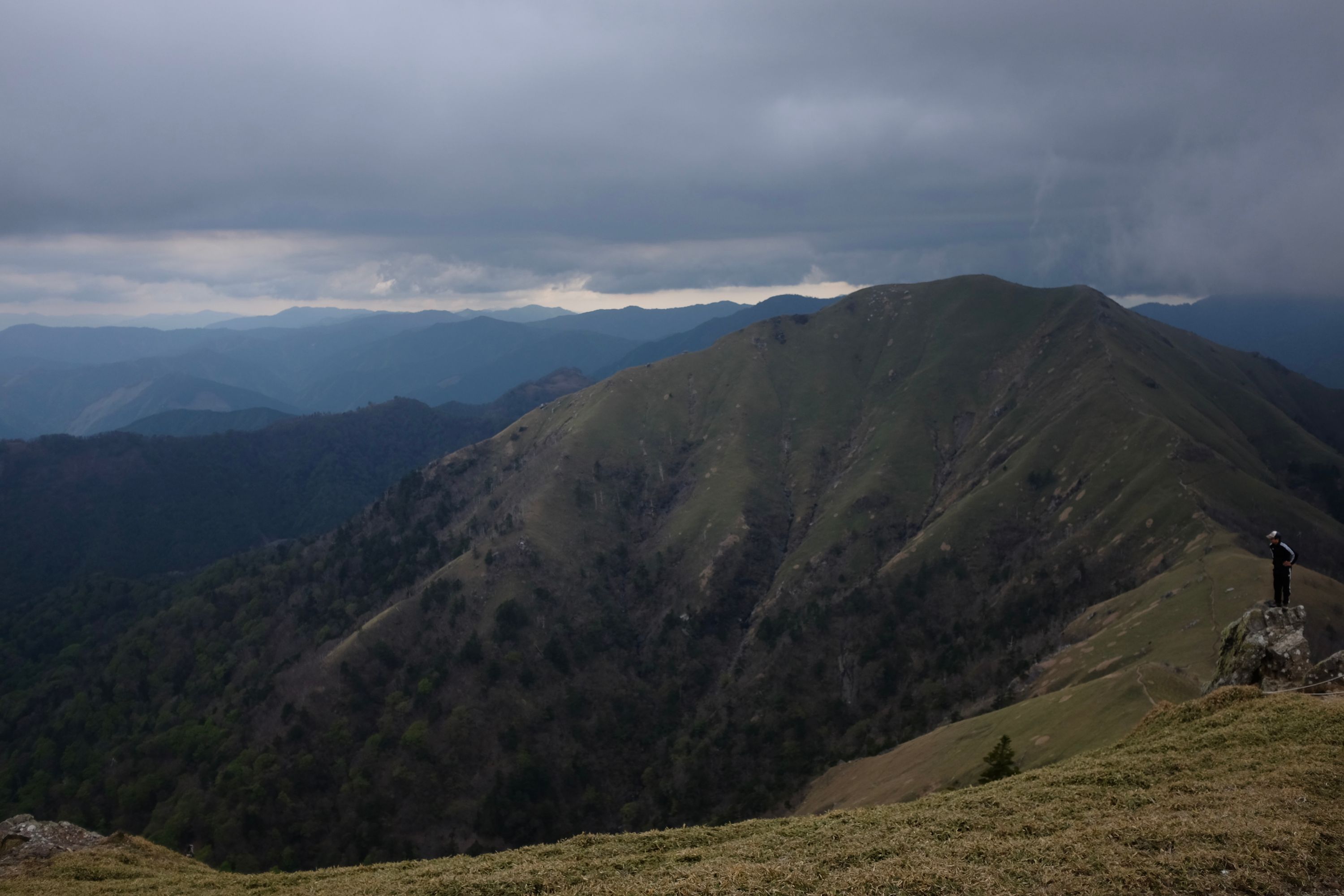 A man stands on a rock with a path along a mountain ridge visible behind him. This is Mount Tsurugi, the second highest point of Shikoku.