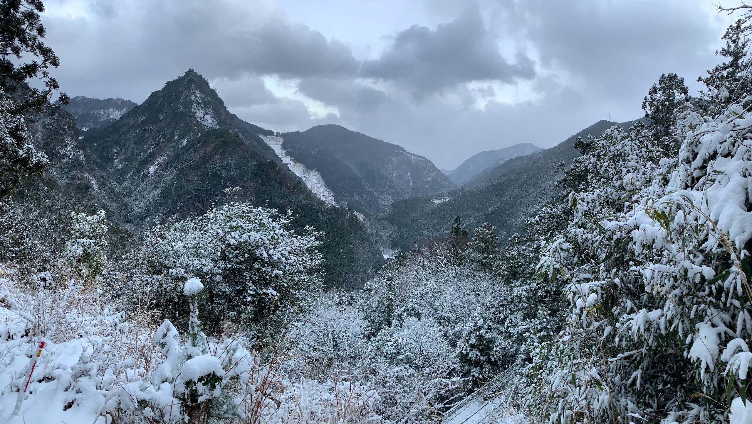 A small but very steep, pyramidal mountain rises from a wintry landscape of snow-covered trees and mountains.